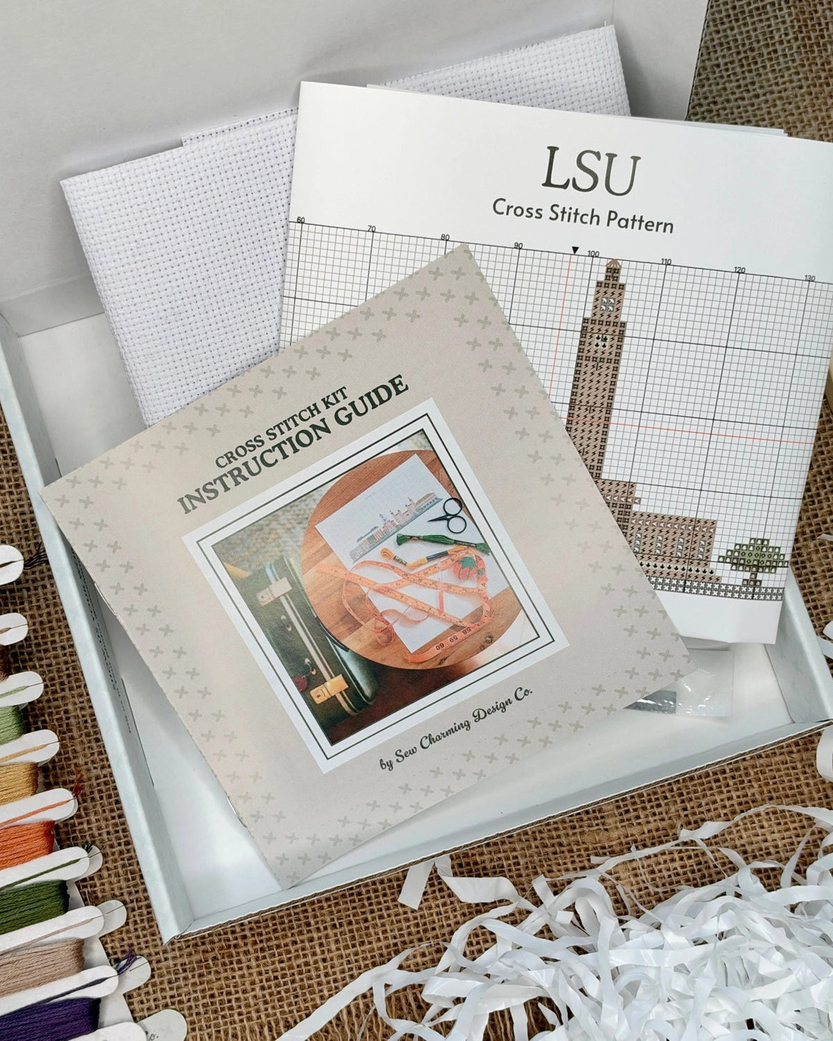 A cross stitch kit opened to reveal a cross stitch pattern booklet, an instruction guide, thread spools in multiple colors, and white thread wrapping paper on a burlap surface.