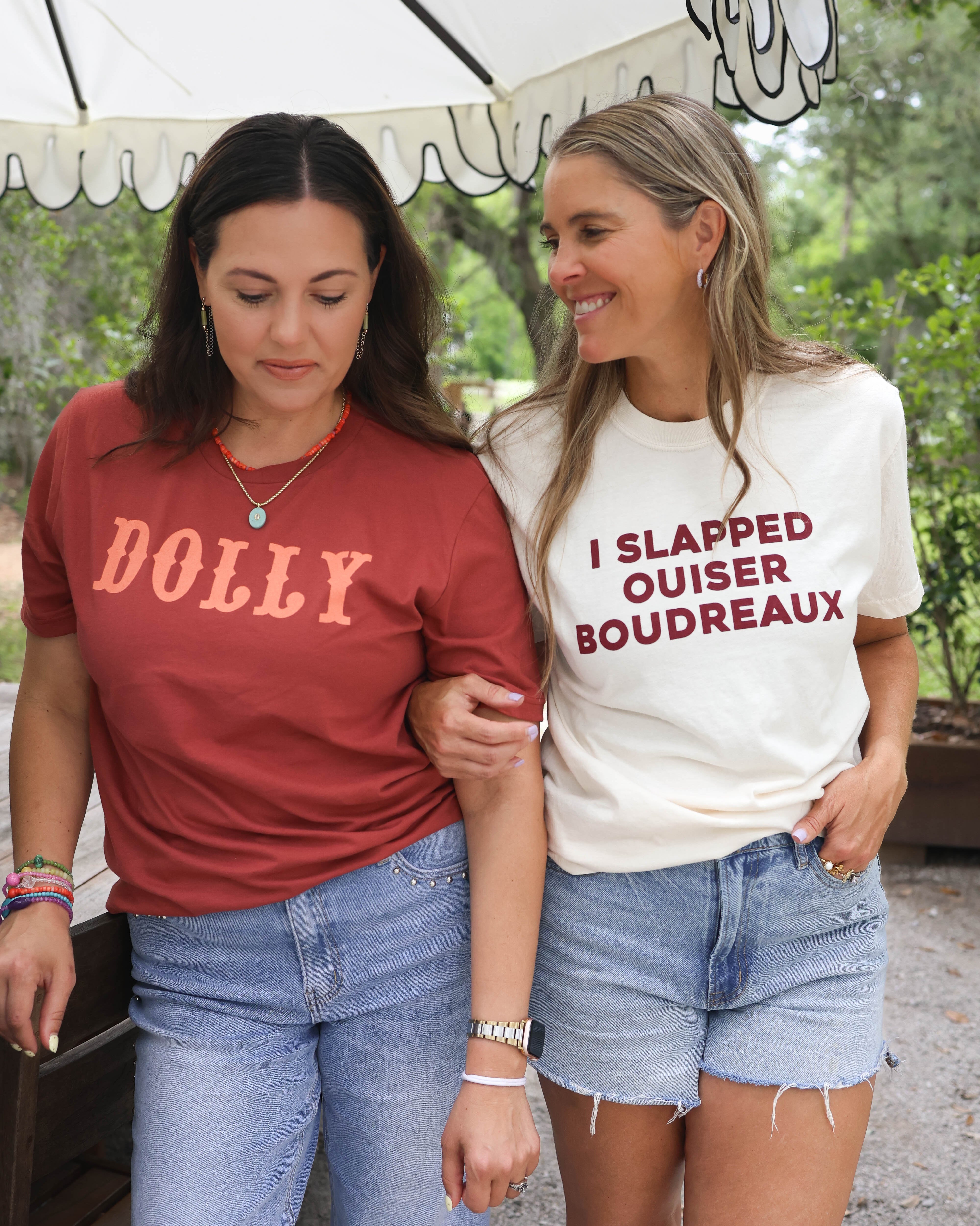 Two women walking outdoors, one wearing a red 'DOLLY' shirt and the other in a white 'I SLAPPED OUISER Boudreaux' shirt.