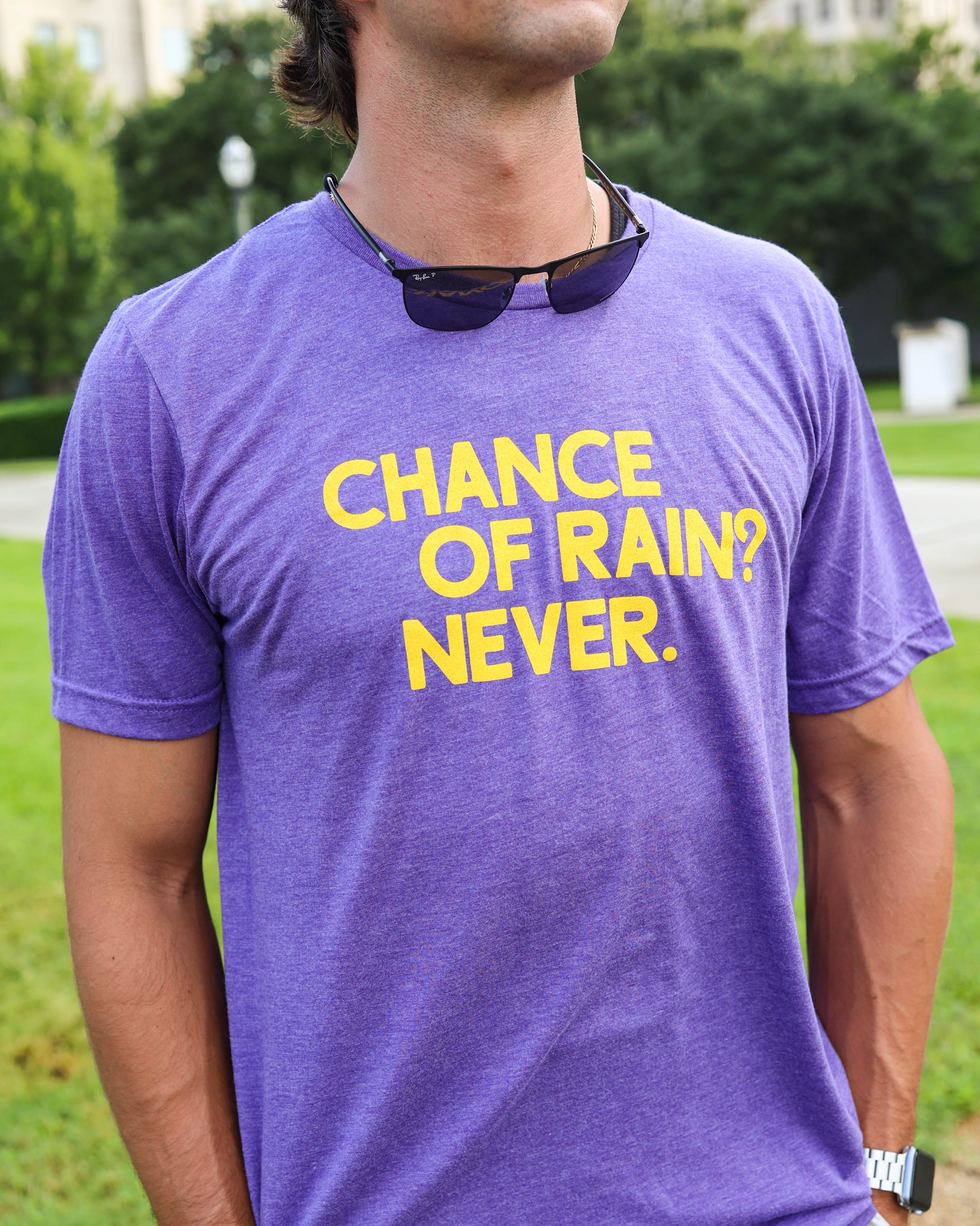 A man wearing a heather purple t-shirt with yellow text that reads 'CHANCE OF RAIN? NEVER.' He is outdoors on a grassy lawn with trees in the background.