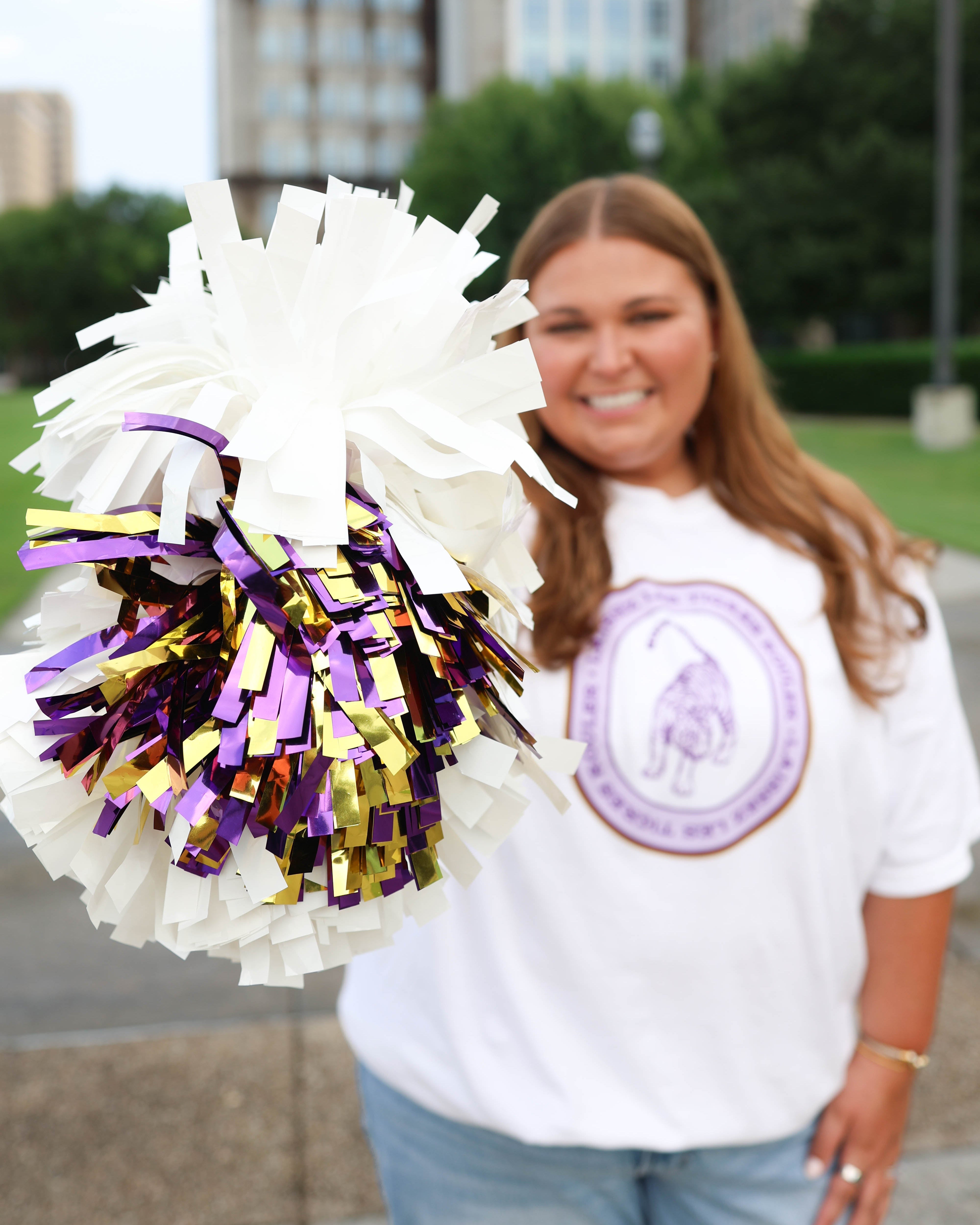 A smiling woman outdoors holding a large white pom-pom with purple and gold tinsel fringe. She is wearing a white graphic t-shirt and blue jeans, with a blurred city background.