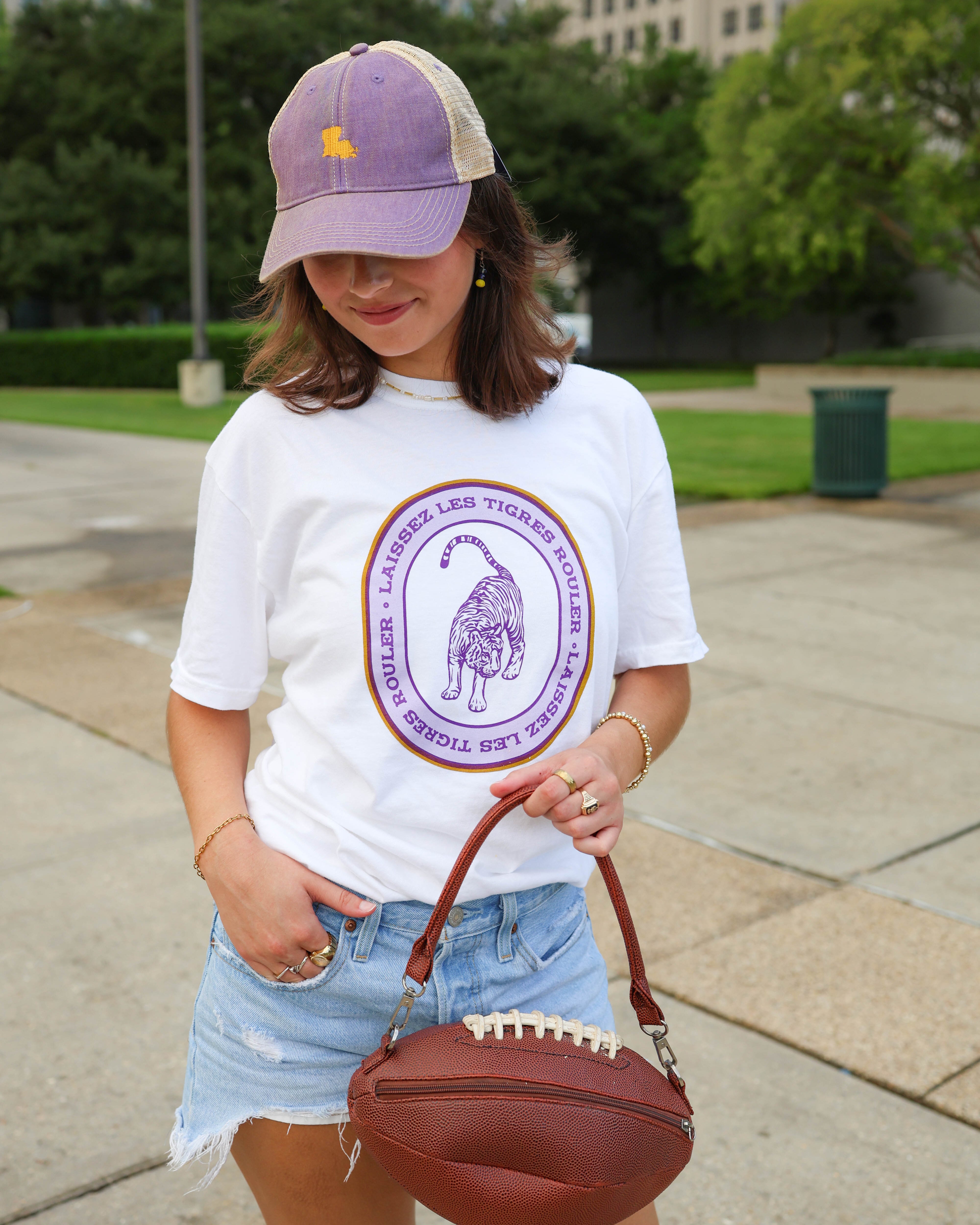 A woman wearing a white graphic t-shirt with a purple circular logo, a purple and beige trucker hat, denim shorts, and holding a brown football-shaped shoulder bag on a paved outdoor walkway.