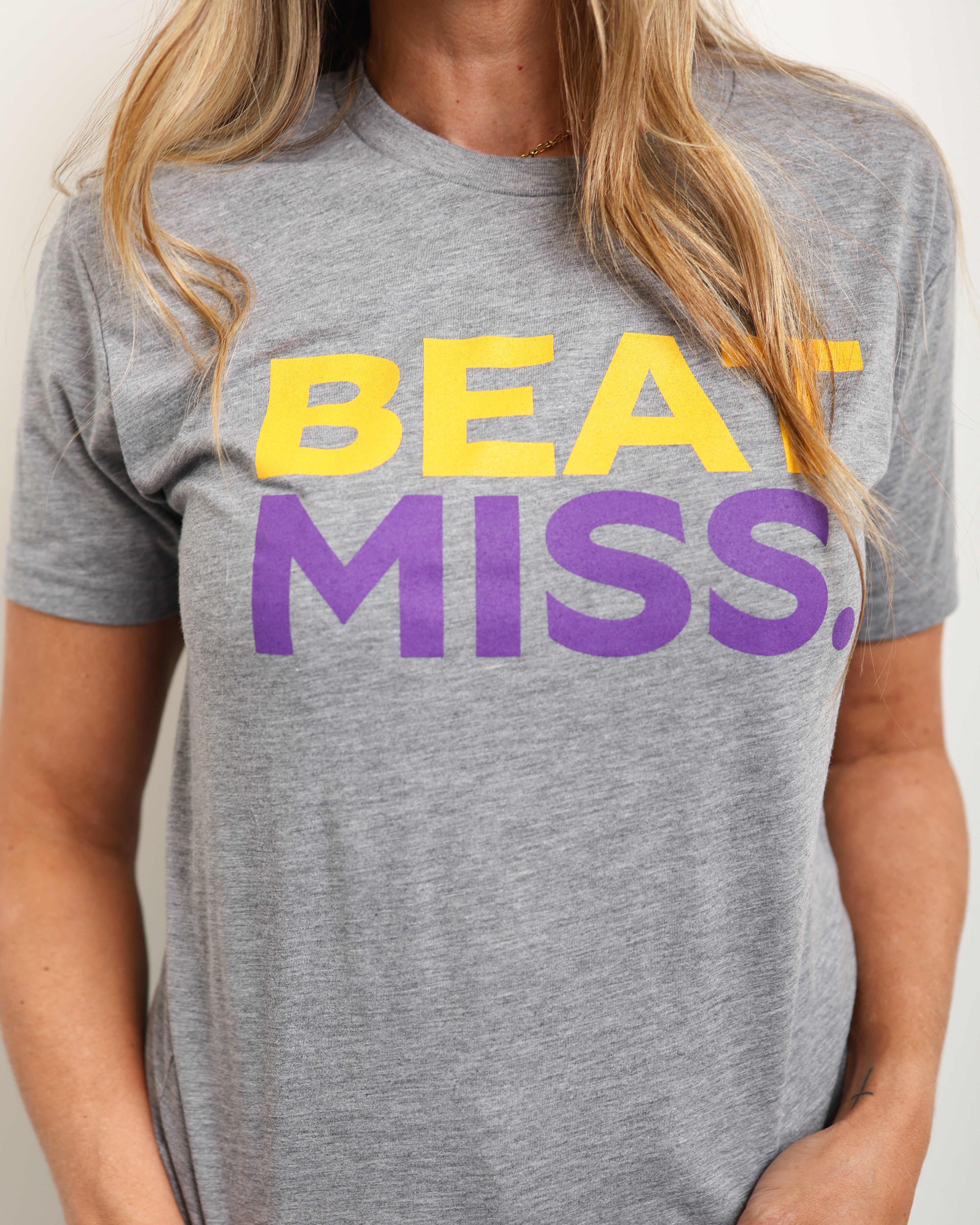 A woman wearing a heather gray t-shirt with the text 'BEAT MISS.' printed across the chest in yellow and purple, paired with blue jeans.