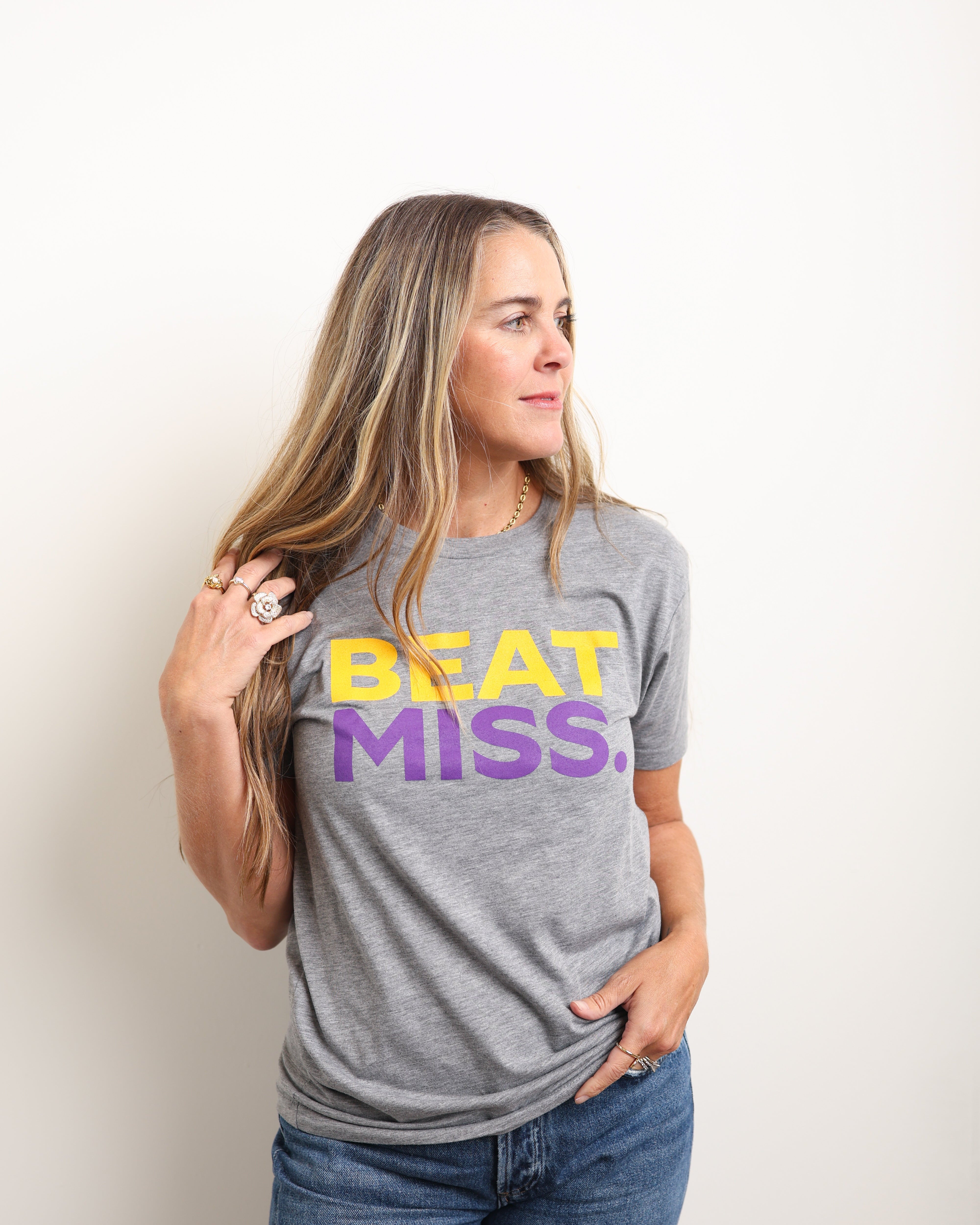 A woman wearing a heather gray graphic t-shirt with 'BEAT MISS.' printed in yellow and purple across the chest, paired with blue jeans, standing against a plain off-white background.
