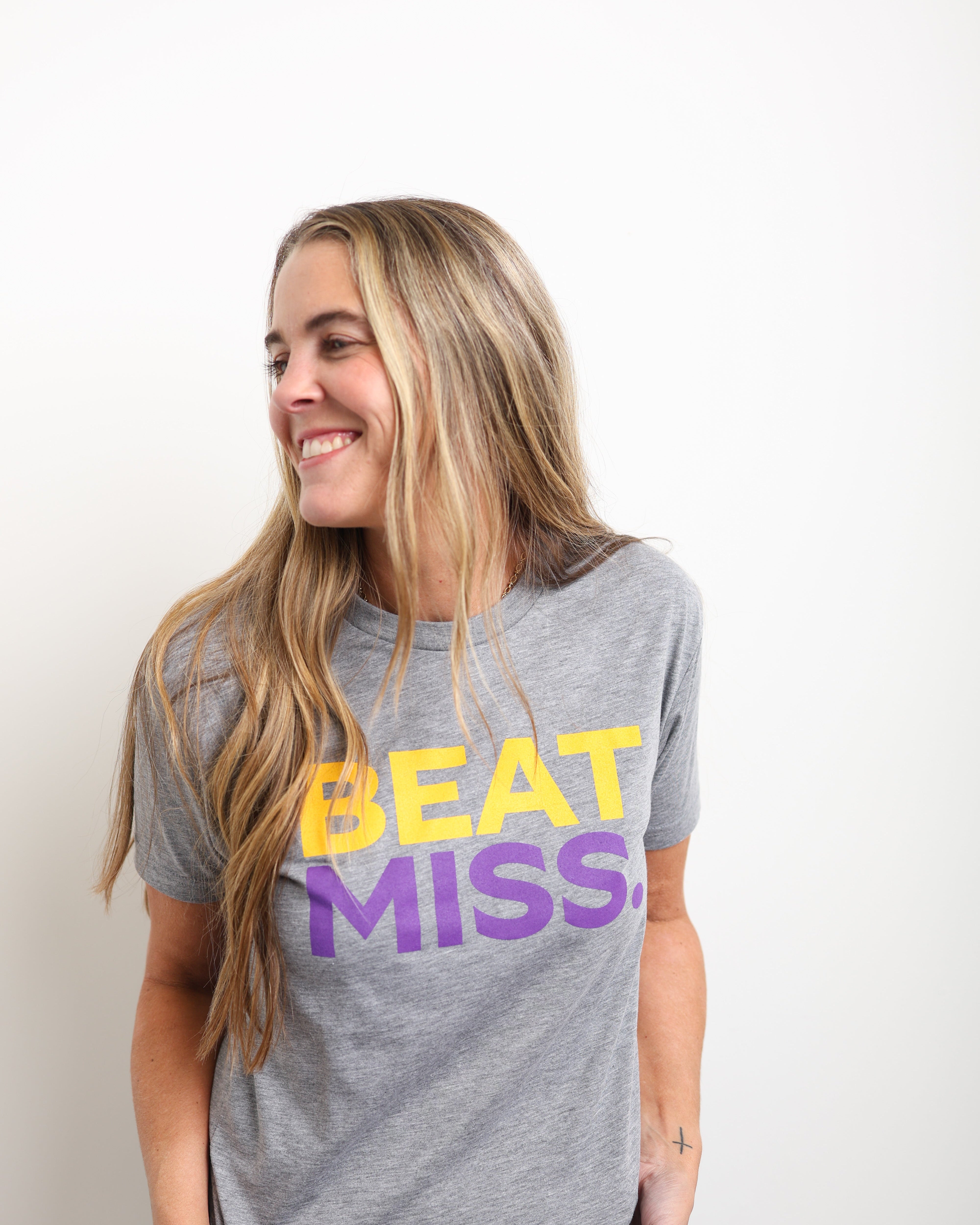 A woman with long blonde hair smiling and looking to the side, wearing a heather gray t-shirt with 'BEAT MISS' printed in yellow and purple text, paired with blue jeans against a plain white background.