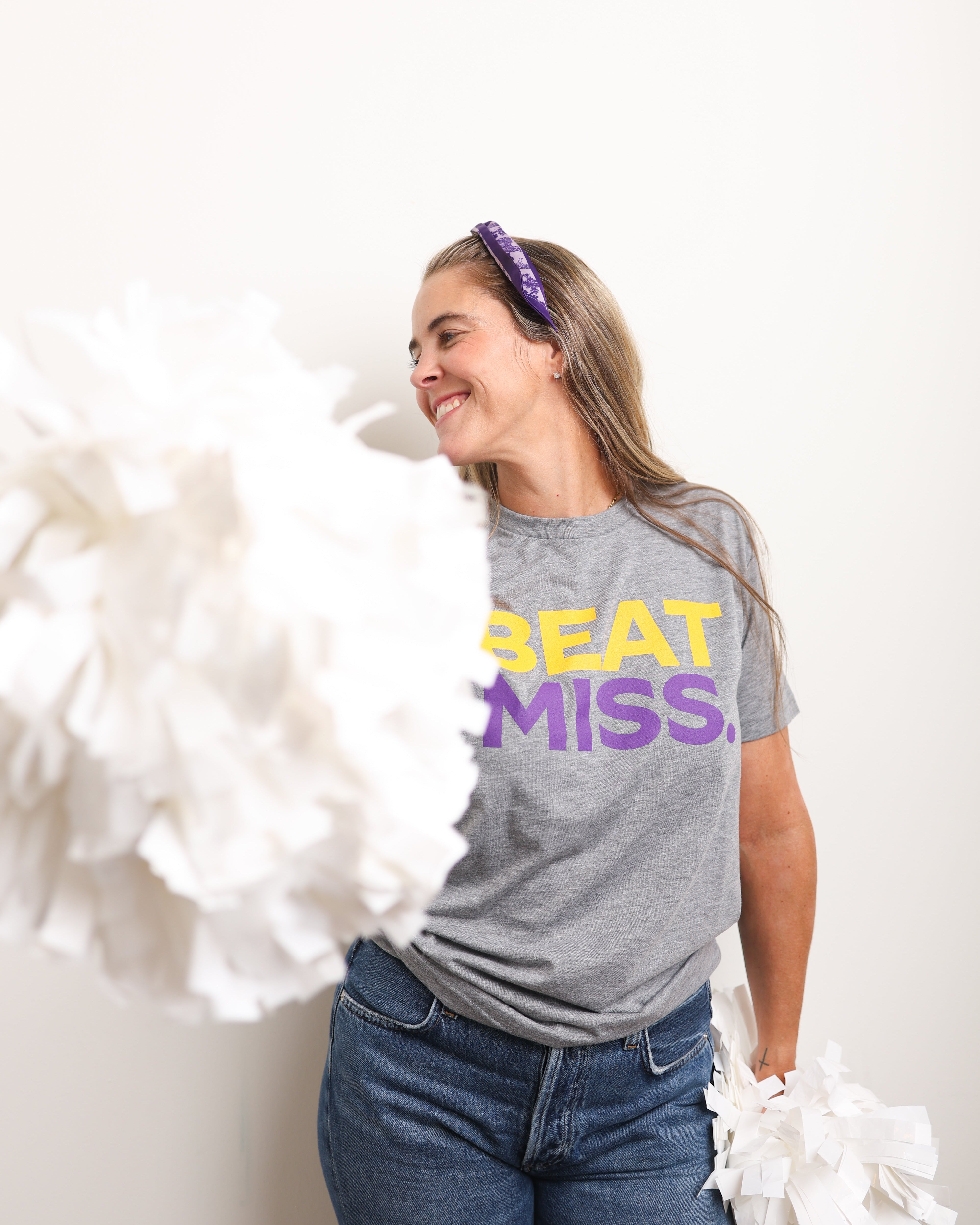 A woman wearing a heather gray graphic t-shirt with 'BEAT MISS.' printed in yellow and purple, paired with blue jeans and a purple headband, holding white pom-poms against a plain white background.