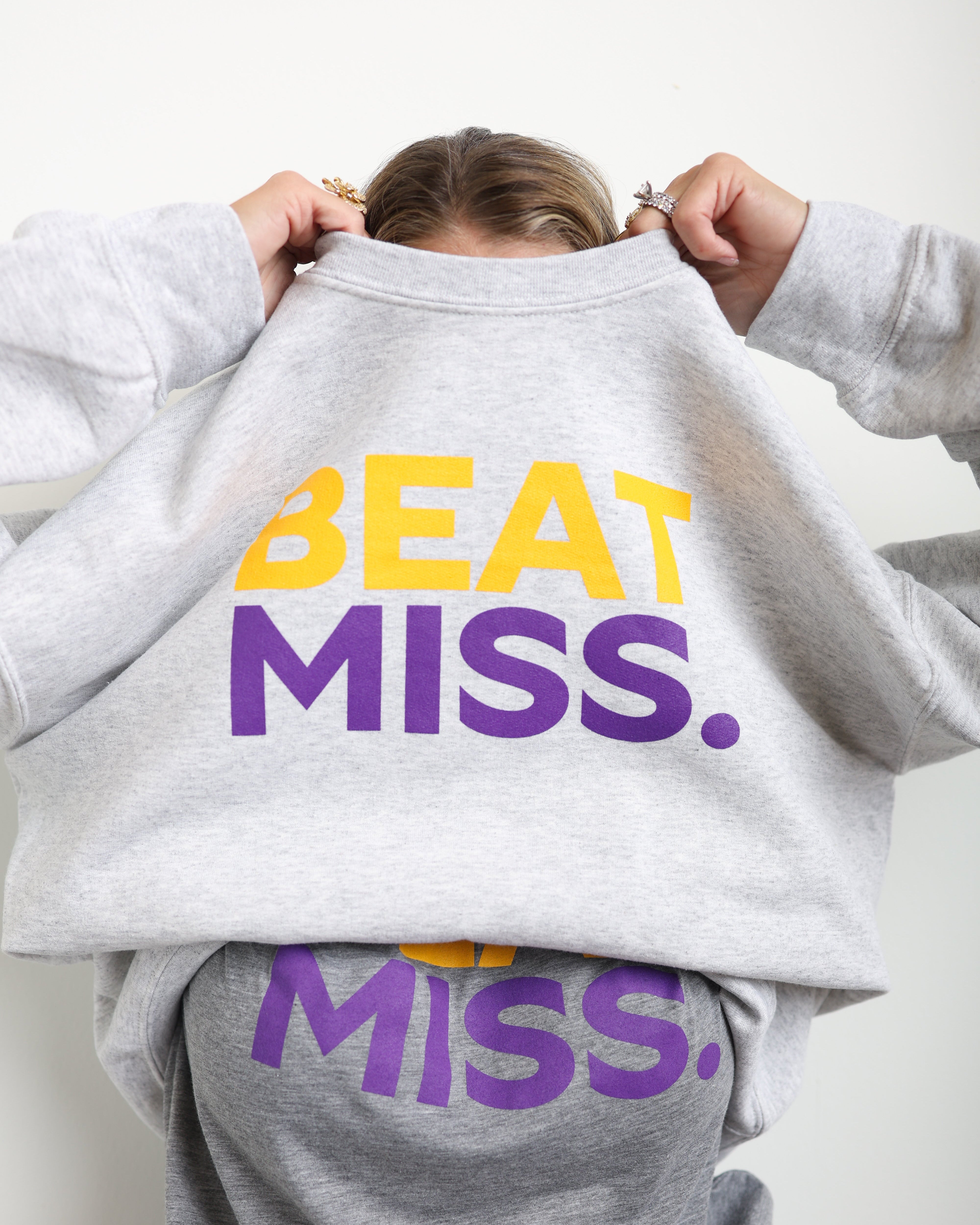 A person wearing a light heather gray sweatshirt with 'BEAT MISS.' printed in yellow and purple across the chest, holding the collar up to show the graphic.