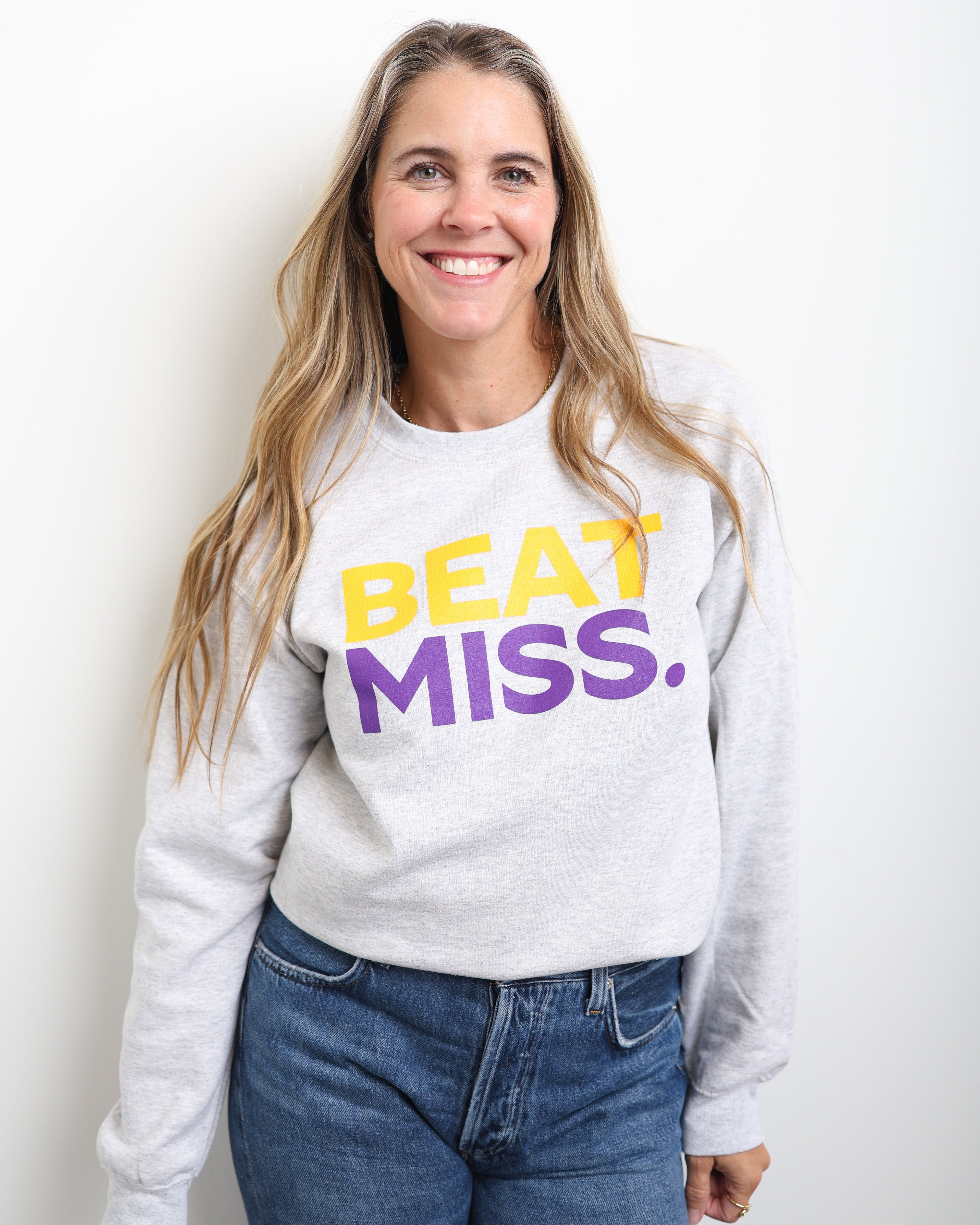 A smiling woman with long blonde hair wearing a light gray sweatshirt with the text 'BEAT MISS.' in yellow and purple, paired with blue jeans, standing against a plain white wall.