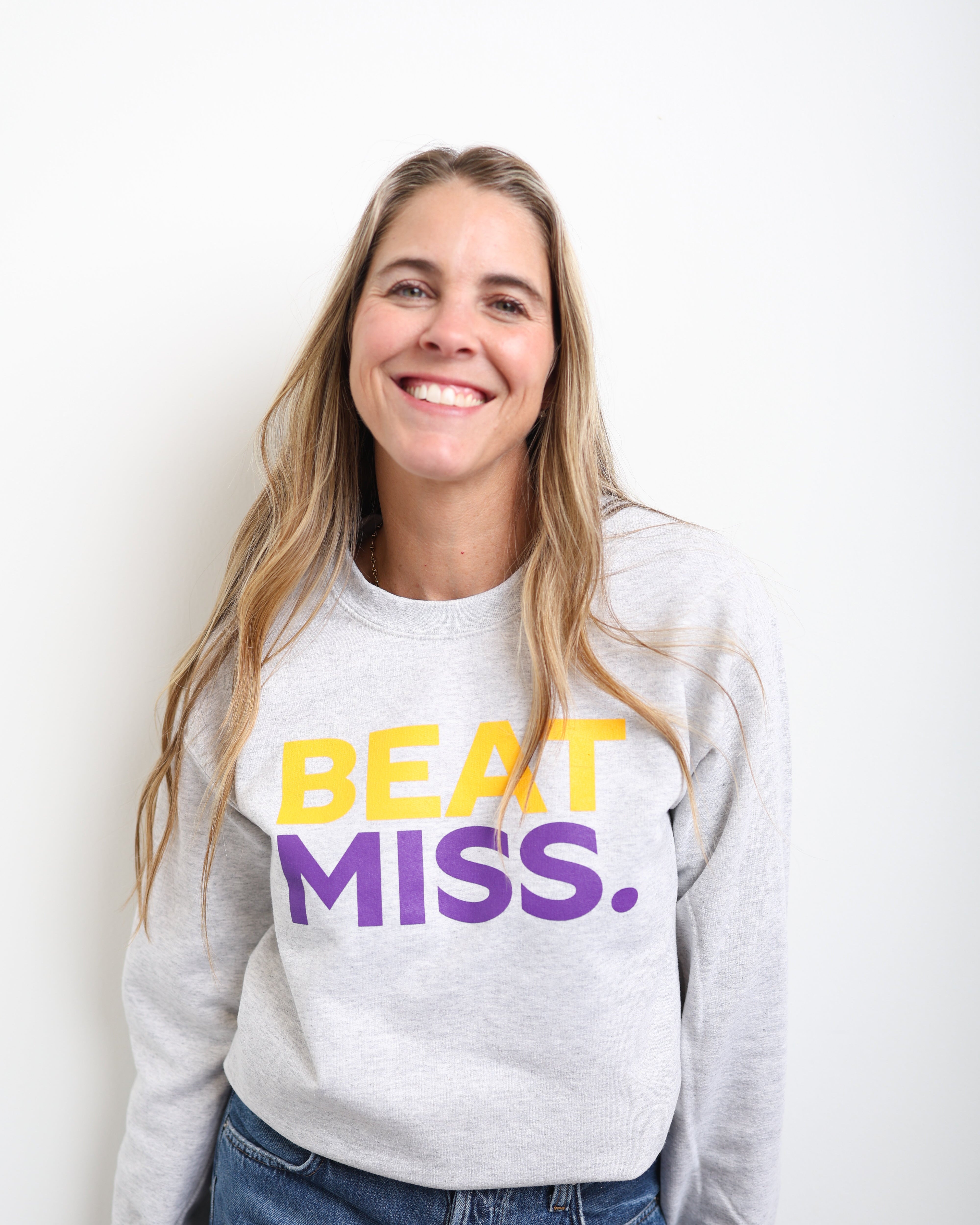 A smiling woman with long blonde hair wearing a light gray sweatshirt with the text 'BEAT MISS.' in yellow and purple, paired with blue jeans, standing against a plain white background.