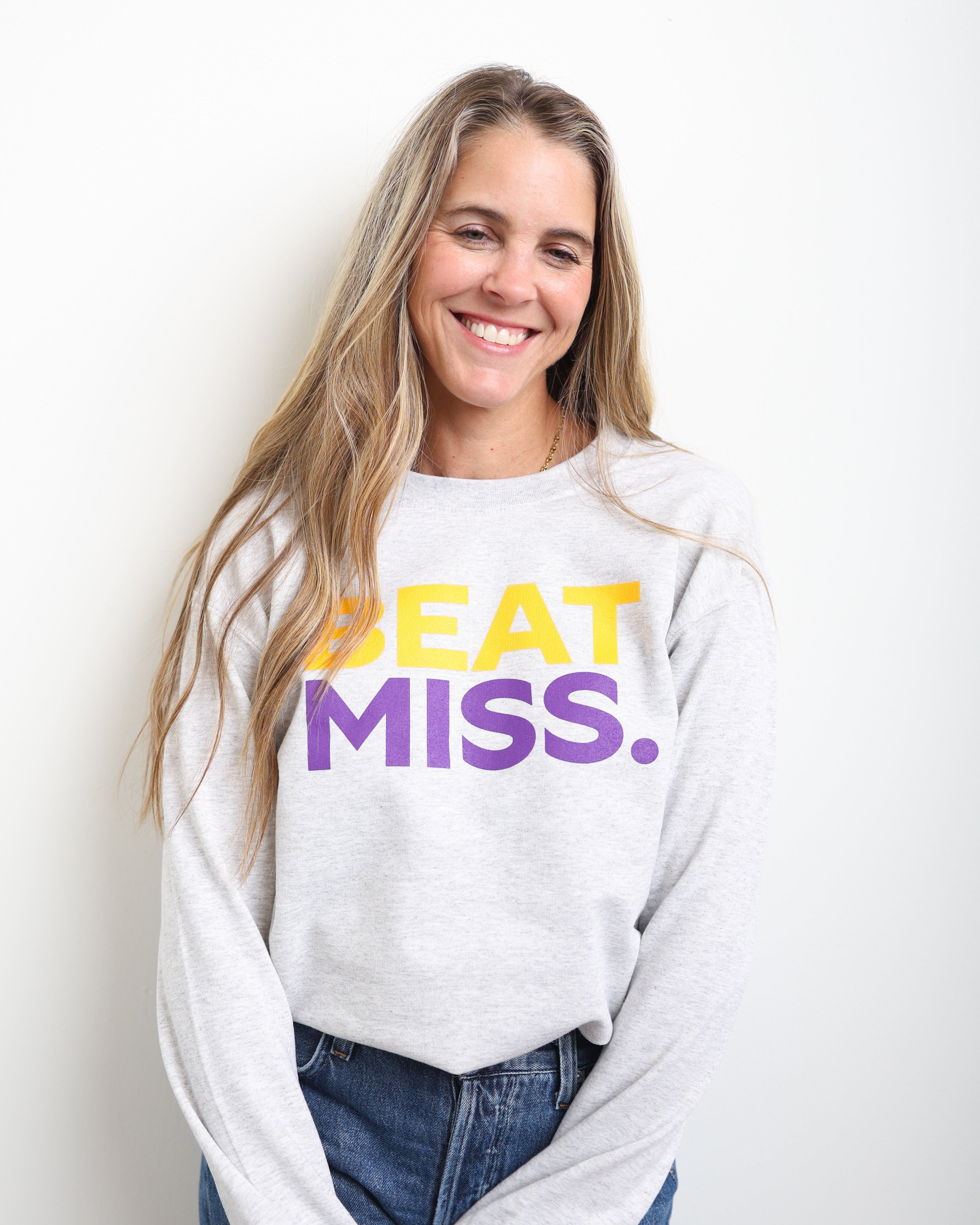 A smiling woman with long blonde hair wears a light gray sweatshirt with the text 'BEAT MISS.' printed on the front in yellow and purple, paired with blue jeans, against a plain white background.