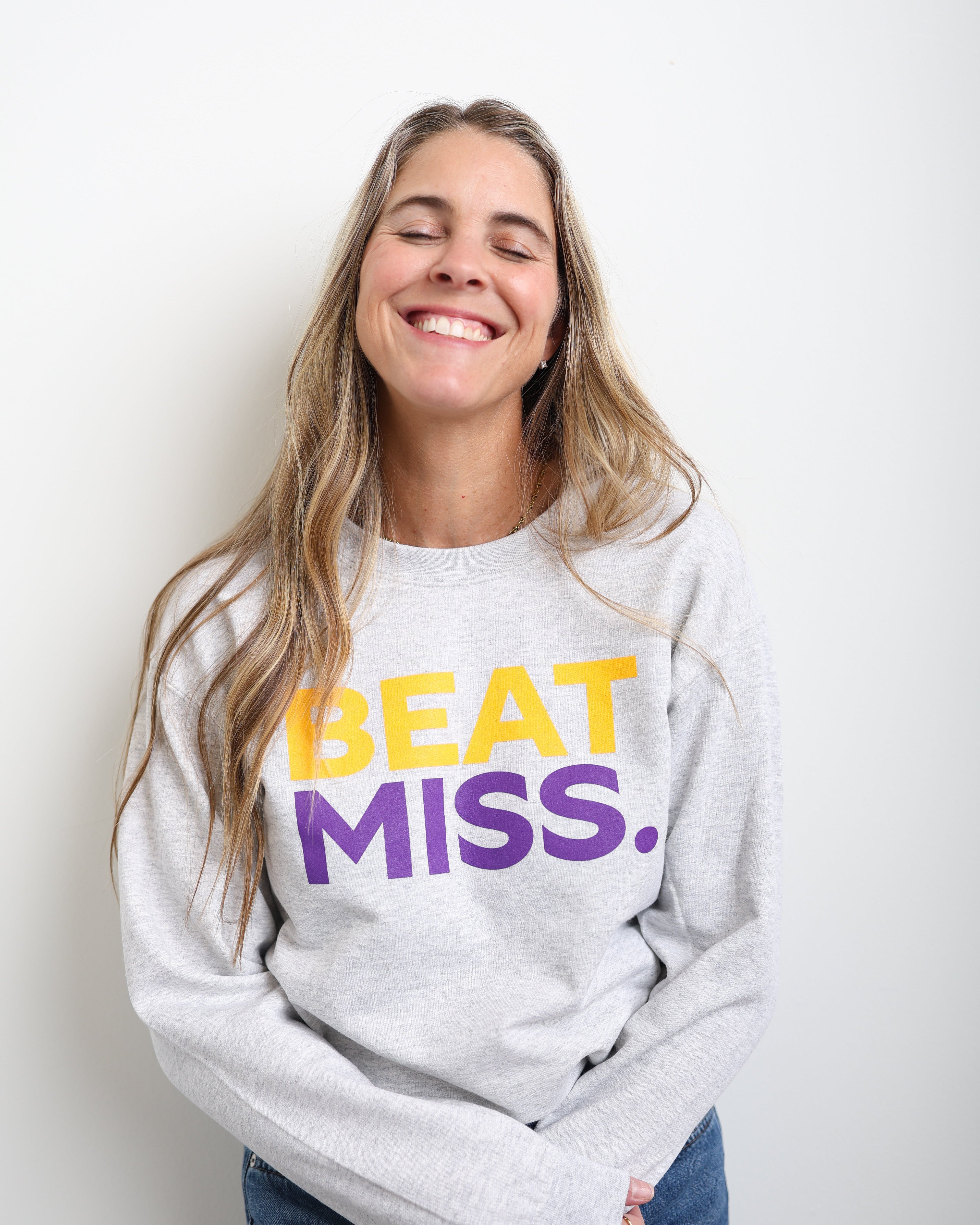 A smiling woman with long blonde hair wearing a light gray sweatshirt with the text 'BEAT MISS.' printed on the front in yellow and purple, paired with blue jeans, against a plain white background.