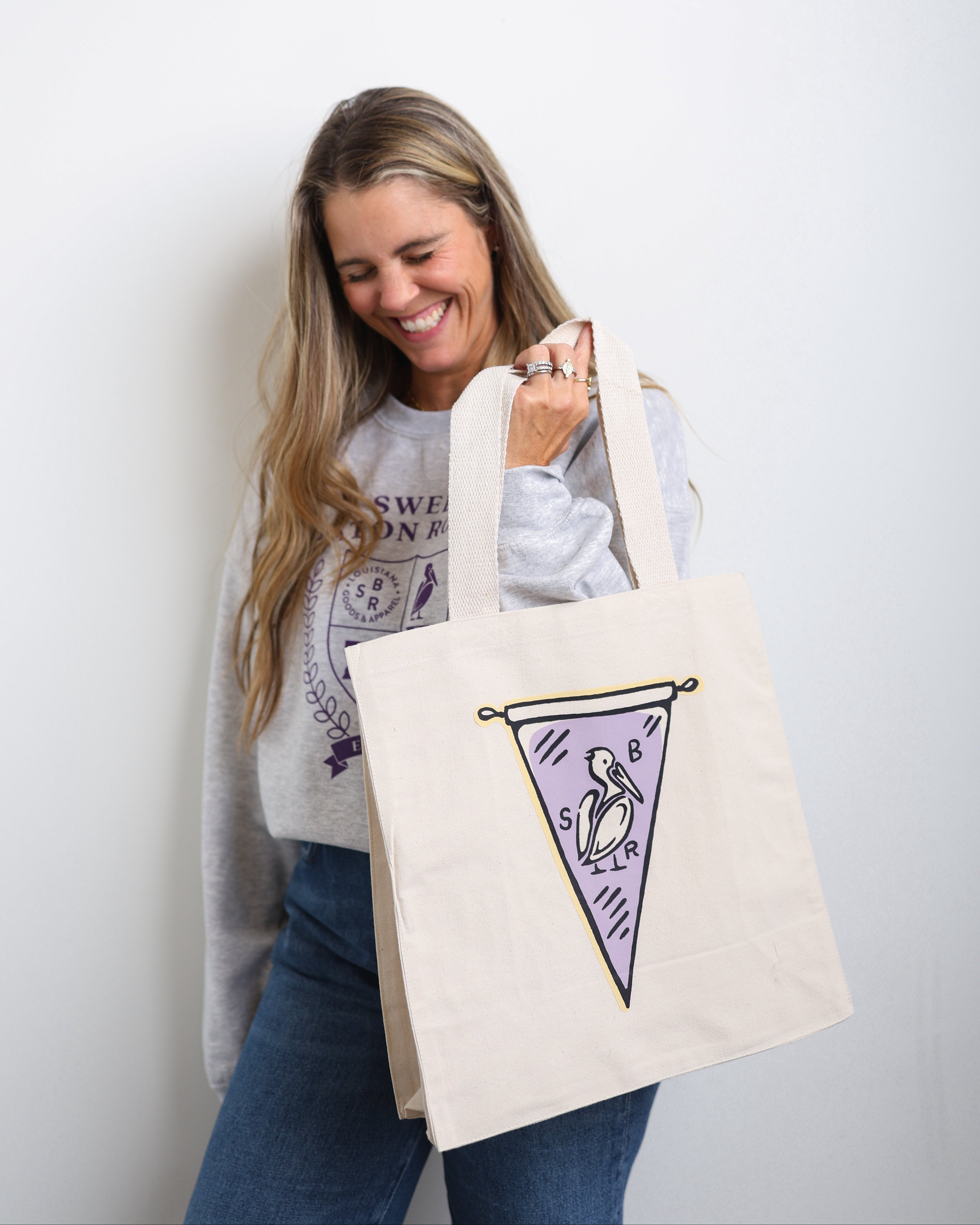 A woman smiling and holding a cream canvas tote bag with purple and black graphics of a pennant flag and a bird. She is wearing a gray graphic sweatshirt and blue jeans against a plain white wall.