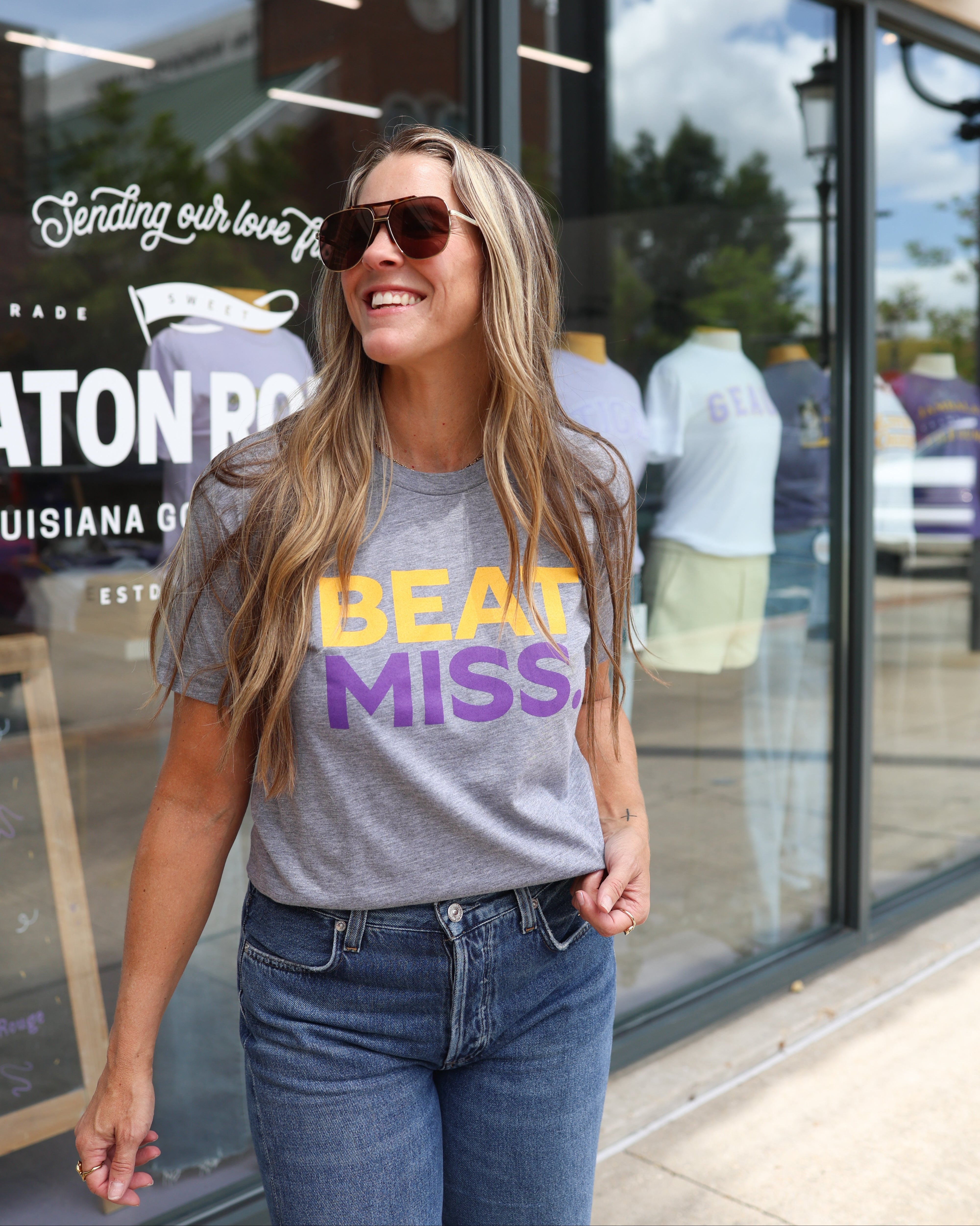 A woman wearing a gray graphic t-shirt with 'BEAT MISS' printed in yellow and purple, paired with blue jeans, standing in front of a storefront window.