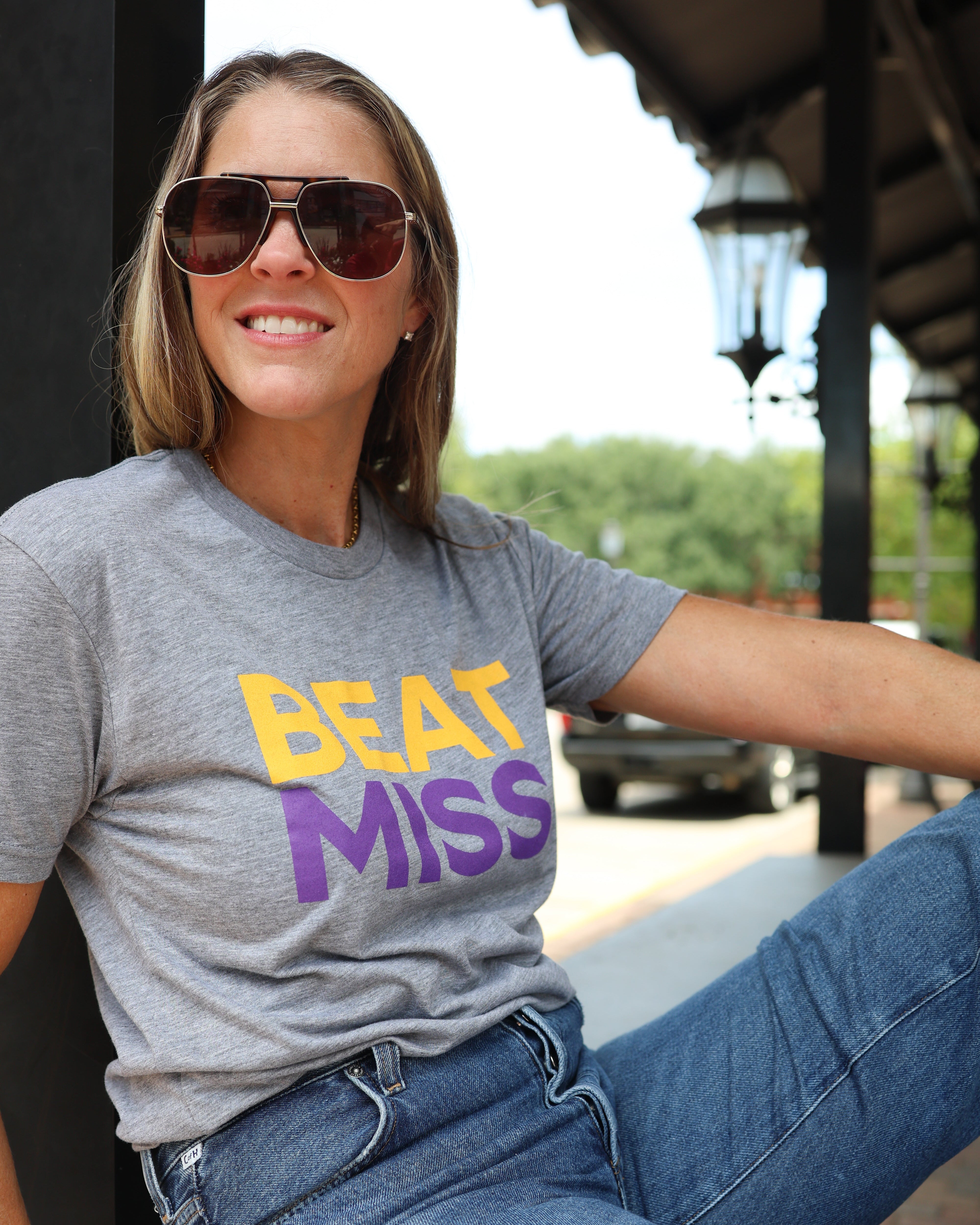 A woman wearing a heather gray graphic t-shirt with 'BEAT MISS' printed in yellow and purple, paired with blue jeans and sunglasses, sitting on an outdoor porch.