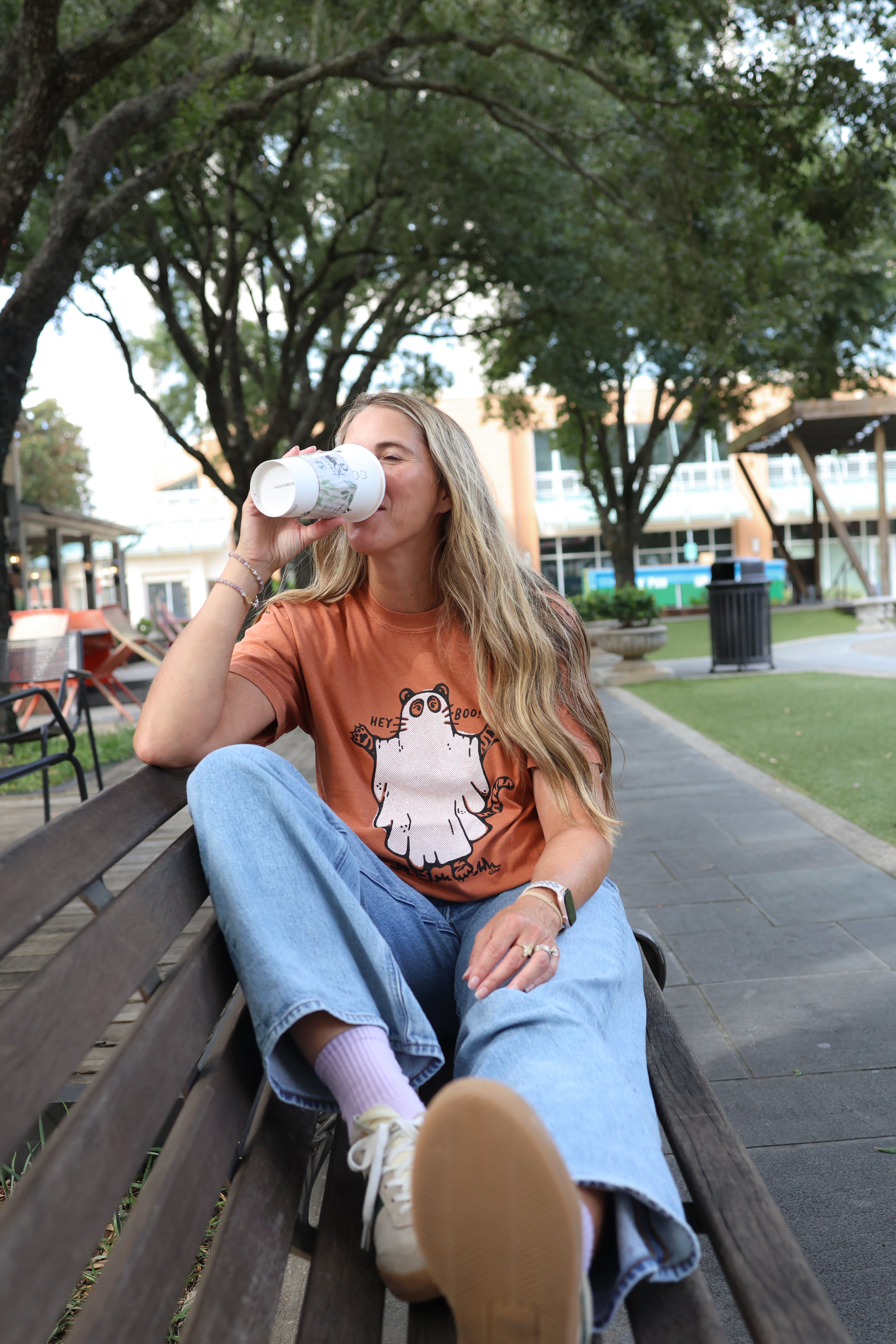 A woman sits on a wooden park bench drinking from a white paper cup, wearing an orange graphic t-shirt, light blue jeans, and white sneakers in an outdoor park setting.