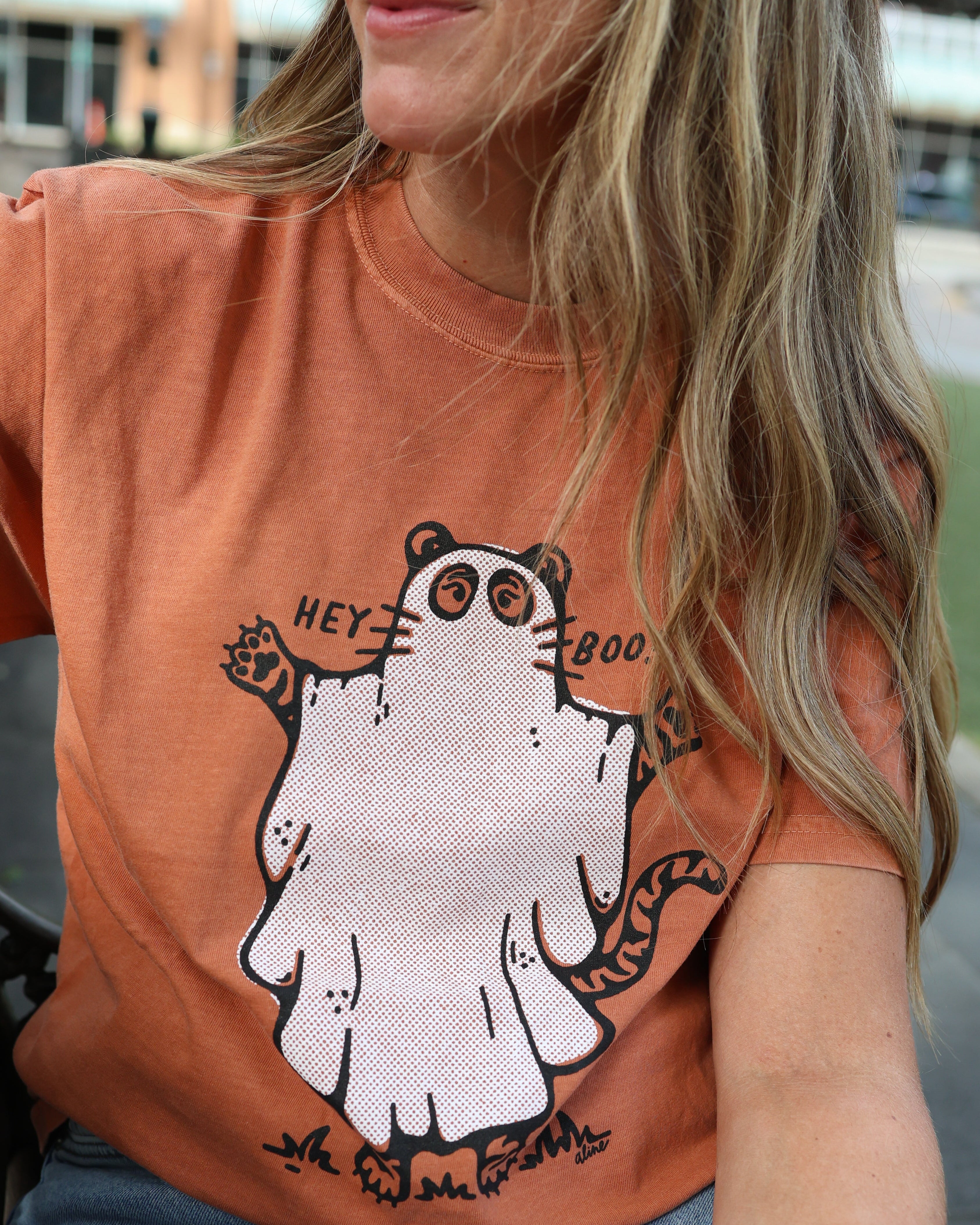 A woman wearing a faded orange crewneck t-shirt with a cartoon tiger dressed as a ghost and the words 'HEY BOO'.