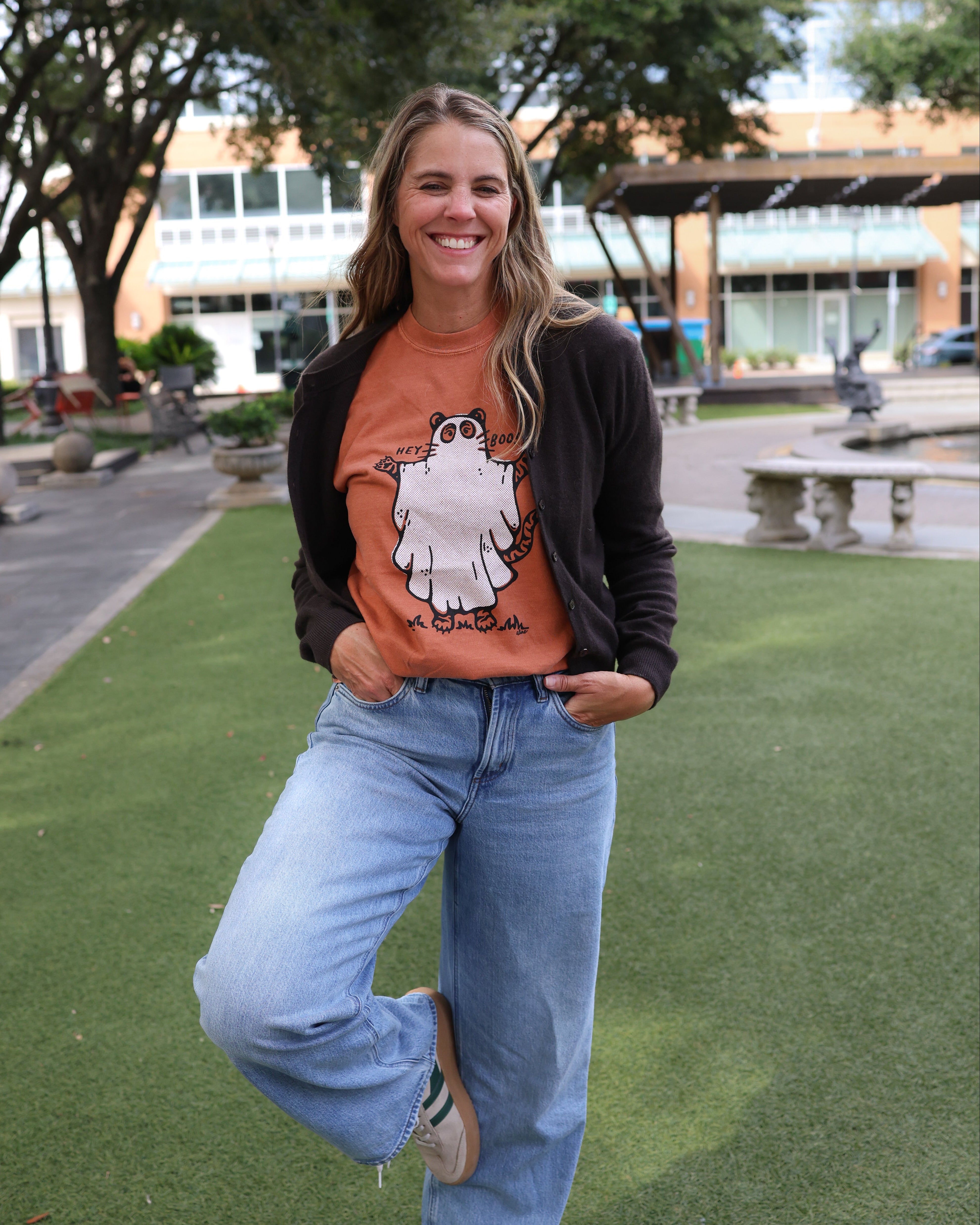 A woman posing outdoors on a green lawn, wearing a black zip-up cardigan over an orange graphic t-shirt with a cartoon ghost, paired with light-wash blue jeans.