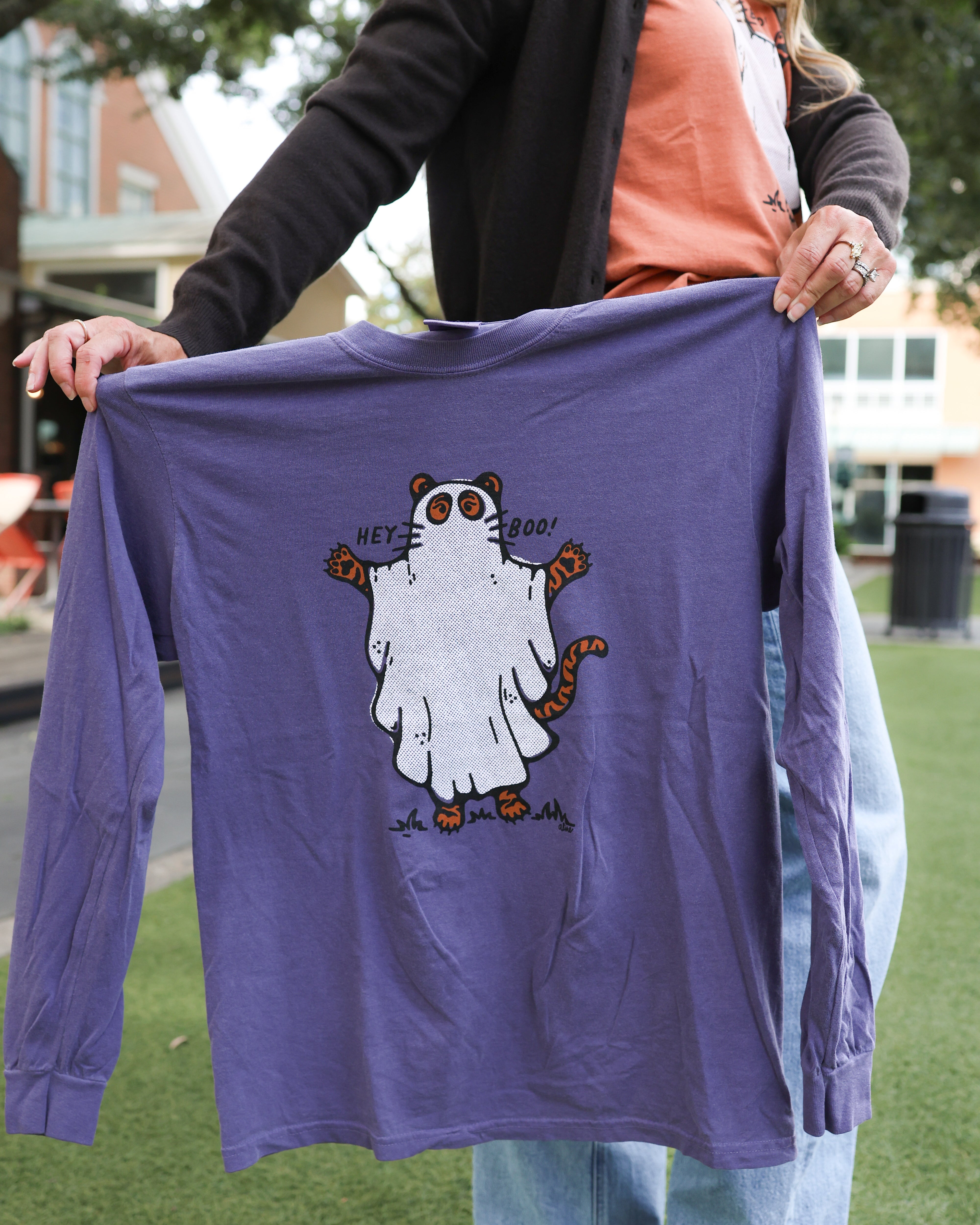 A person outdoors holding up a purple long-sleeve t-shirt with a cartoon ghost tiger graphic on the front.