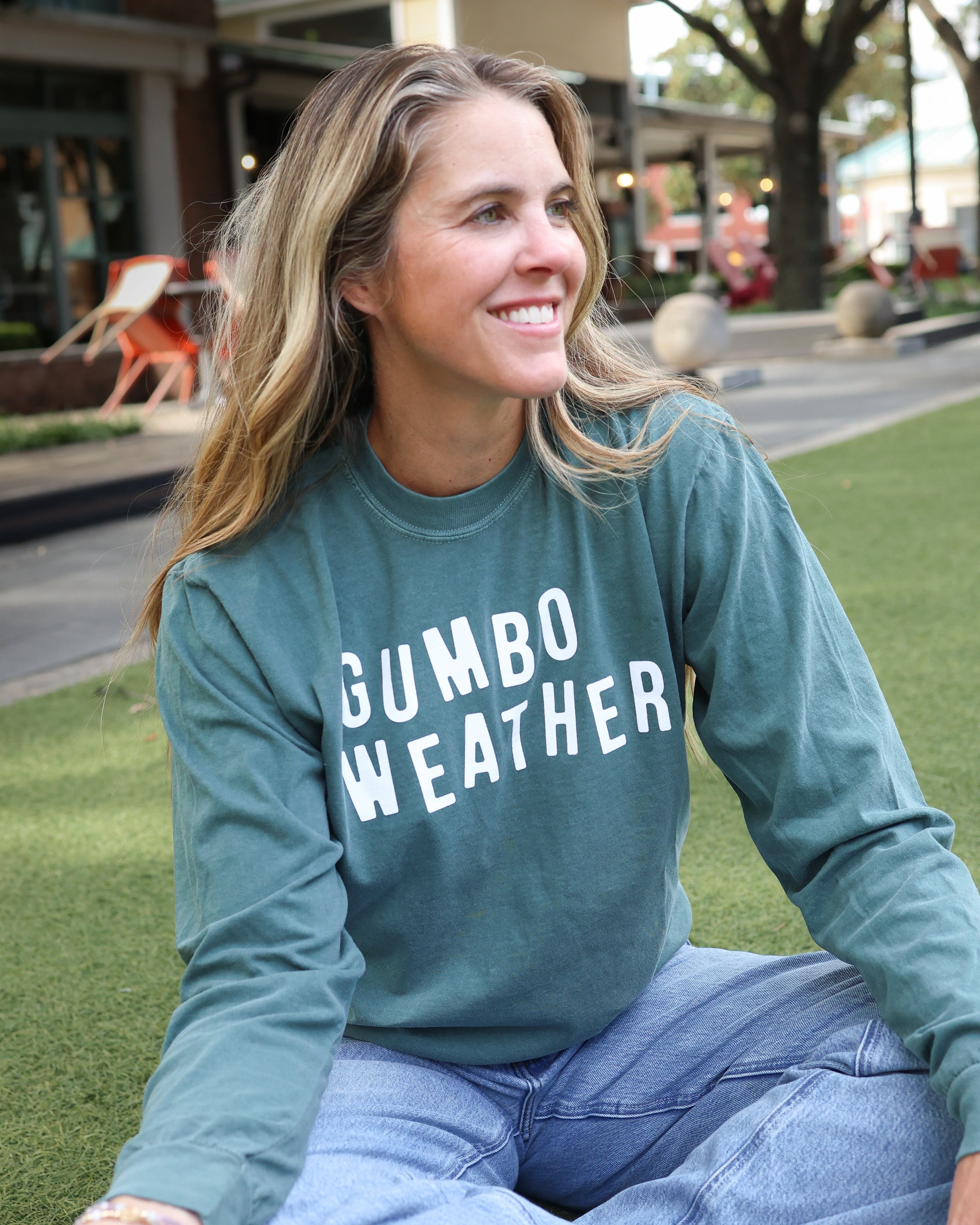 A woman sitting on green grass outdoors wearing a sage green long-sleeve crewneck sweatshirt with 'GUMBO WEATHER' printed in white across the chest, paired with light blue jeans.
