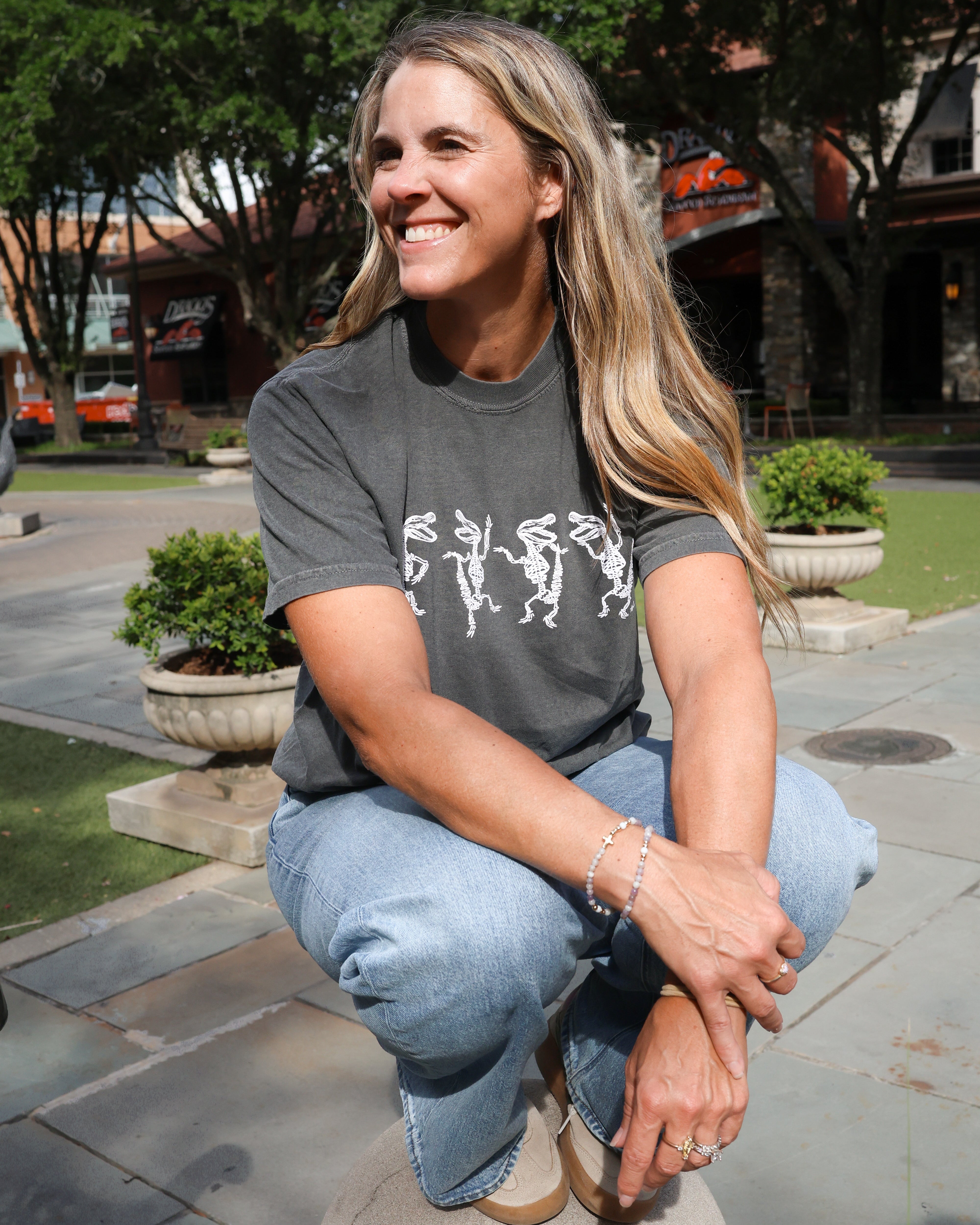 A woman with blonde hair crouching outdoors on a stone pedestal, wearing a dark gray graphic t-shirt and light blue jeans.