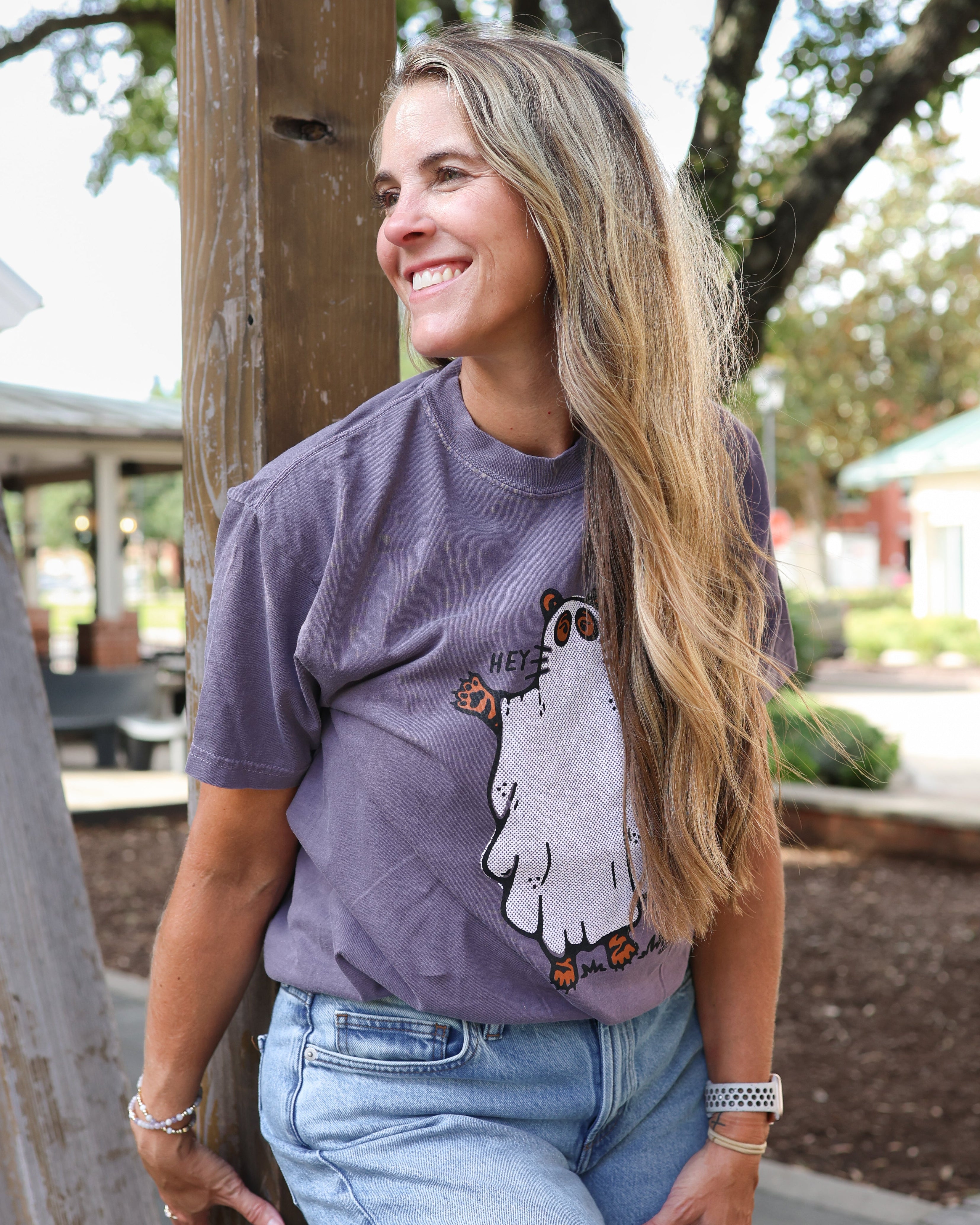 A woman with long blonde hair leans against a wooden post outdoors, wearing a purple graphic t-shirt with a cartoon ghost design and light blue jeans.