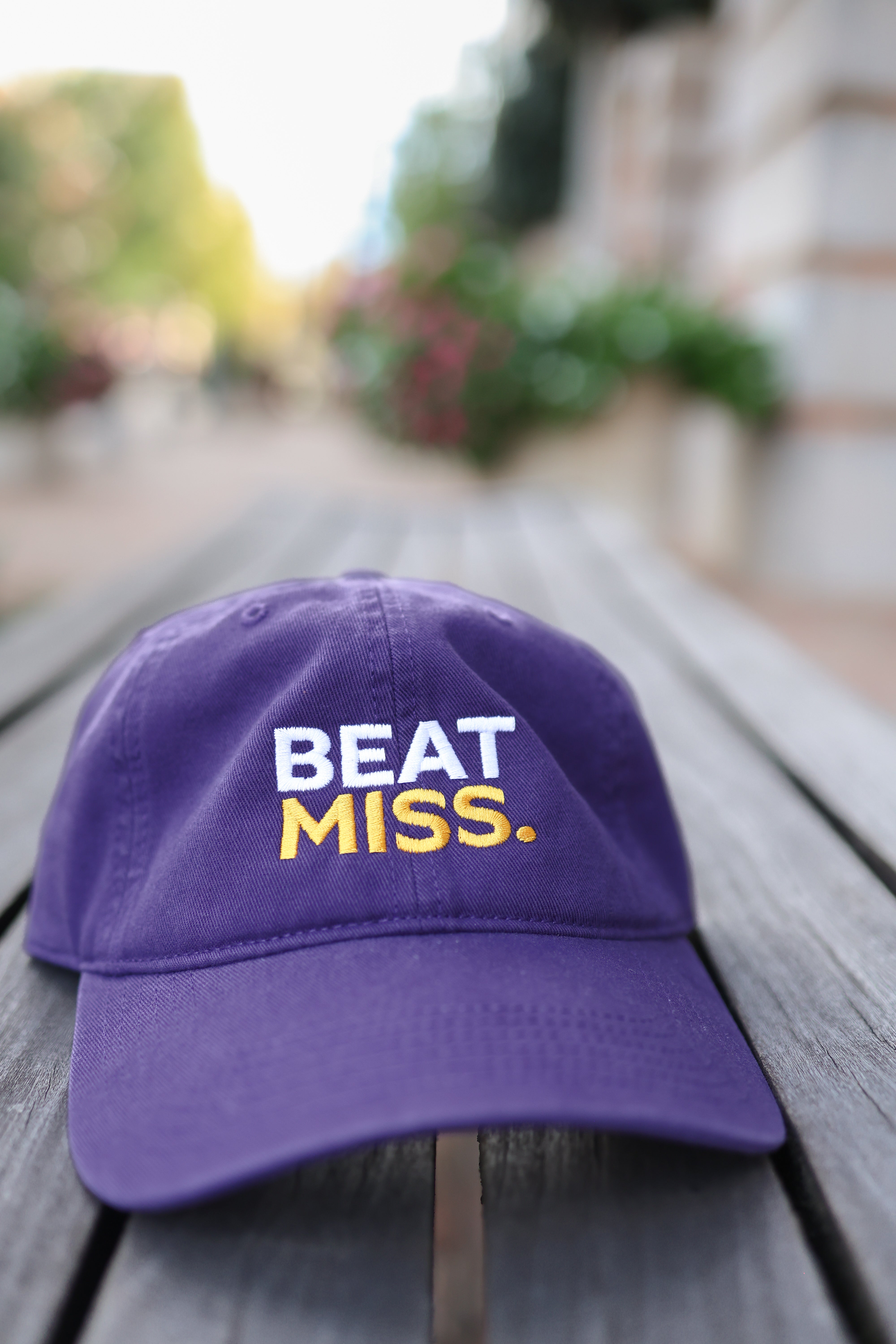 A purple baseball cap with 'BEAT MISS.' embroidered on the front, resting on a weathered wooden bench with a blurred outdoor background.