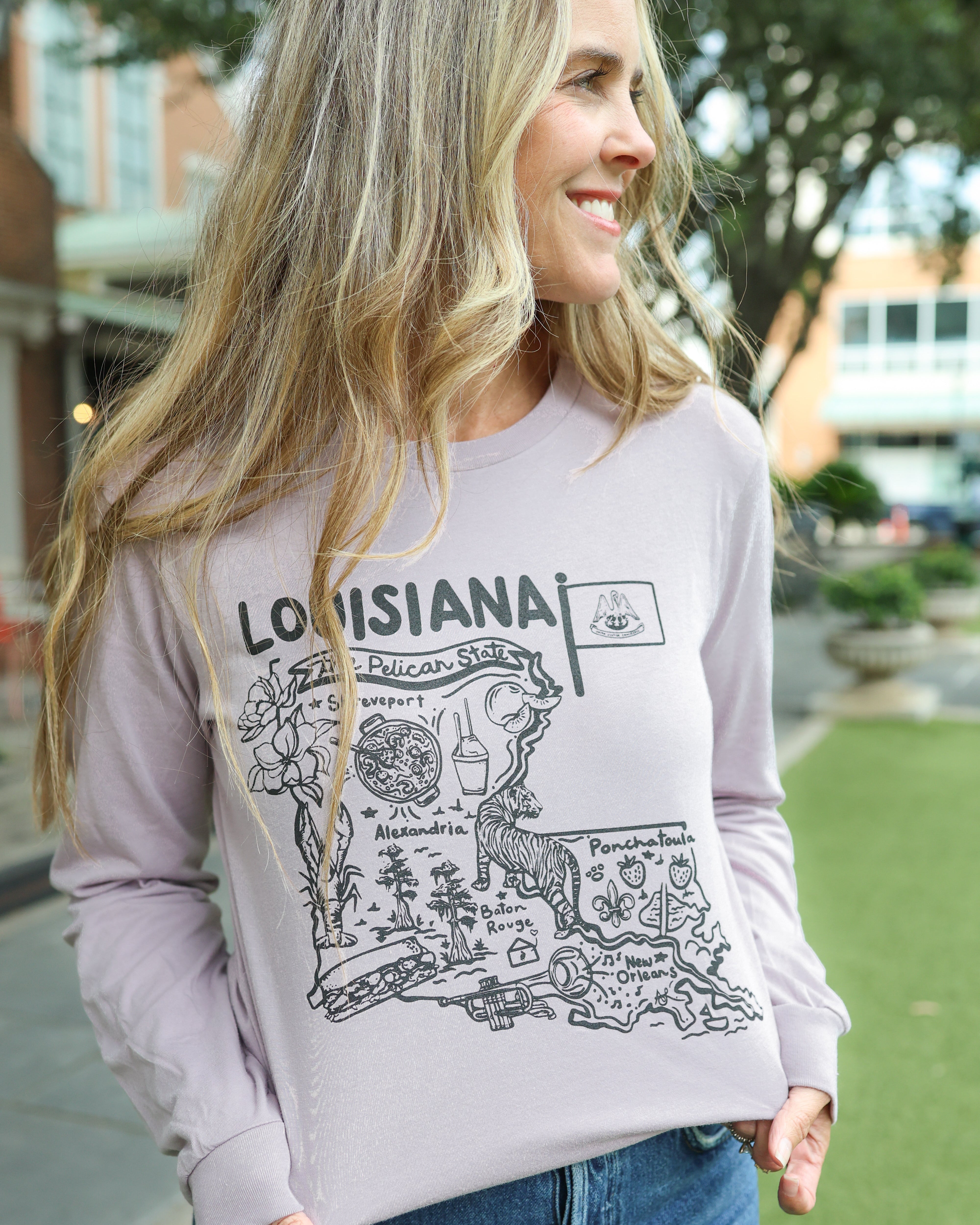 A woman wearing a light lavender crewneck sweatshirt with a black Louisiana state map graphic and 'LOUISIANA' text, paired with blue jeans, standing outdoors on a grassy lawn.