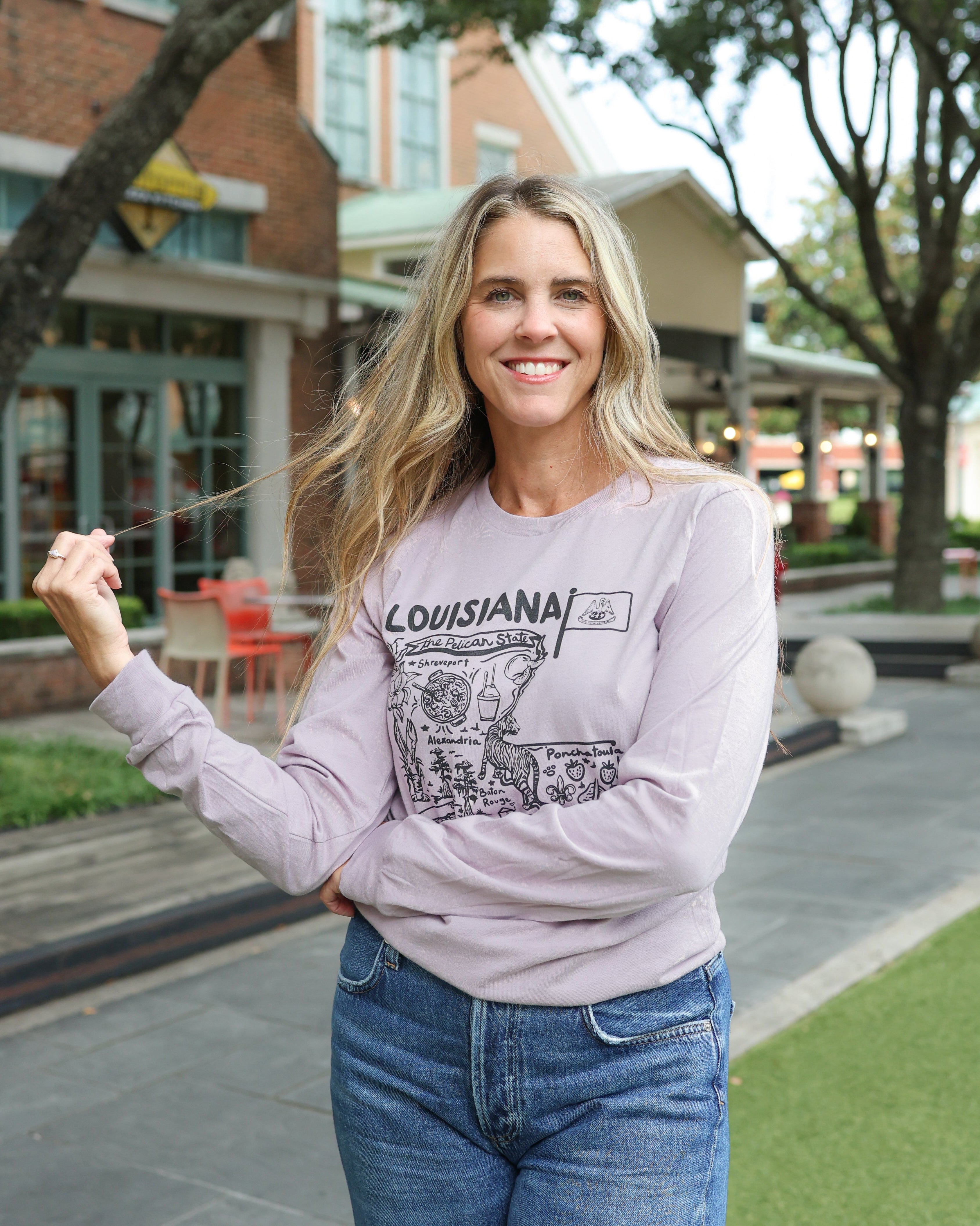 A woman smiling outdoors wearing a light lavender long-sleeve graphic sweatshirt with 'LOUISIANA' and a map design, paired with blue jeans.