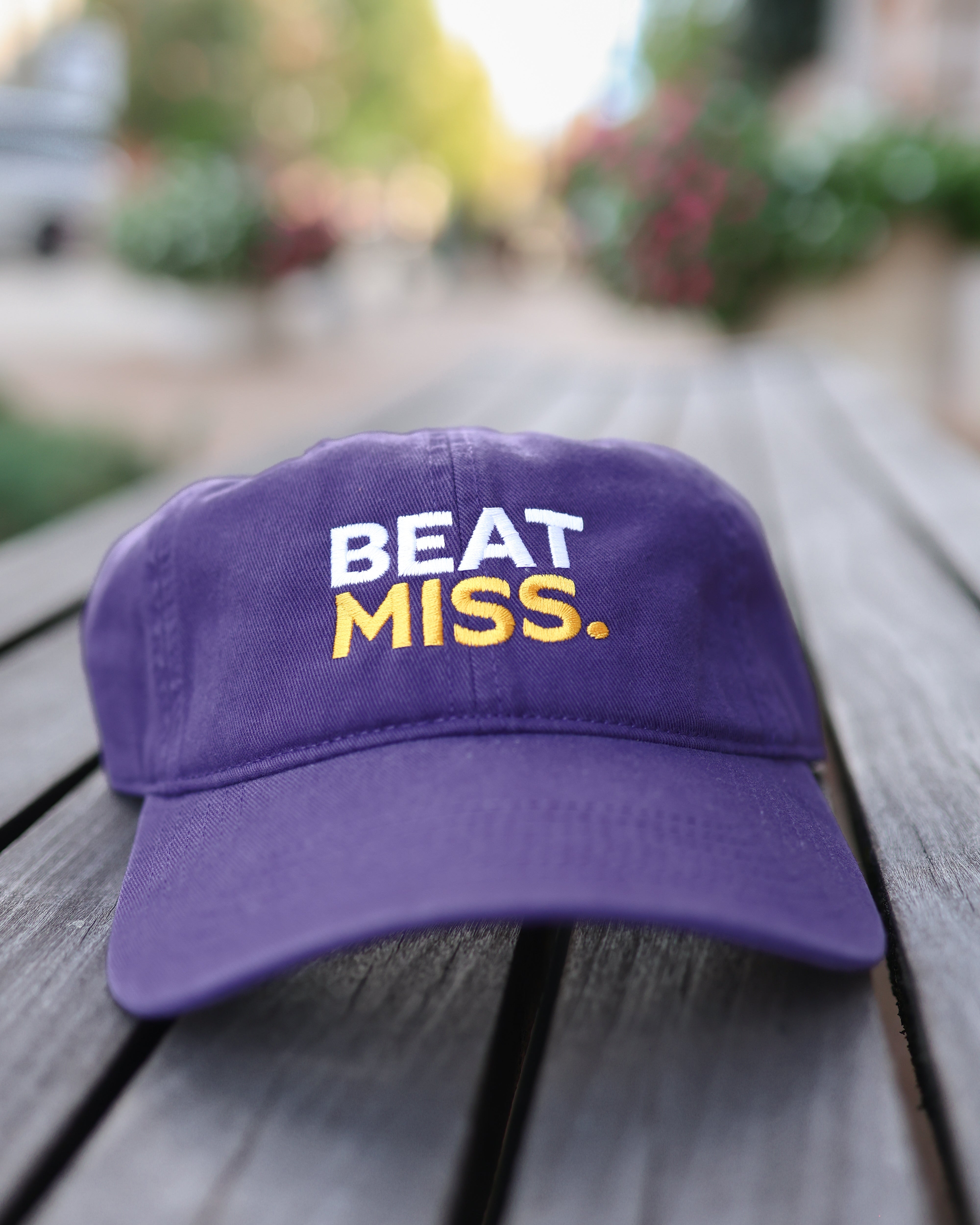 A purple baseball cap resting on a weathered wooden table, featuring the embroidered text 'BEAT MISS.' in white and yellow on the front.