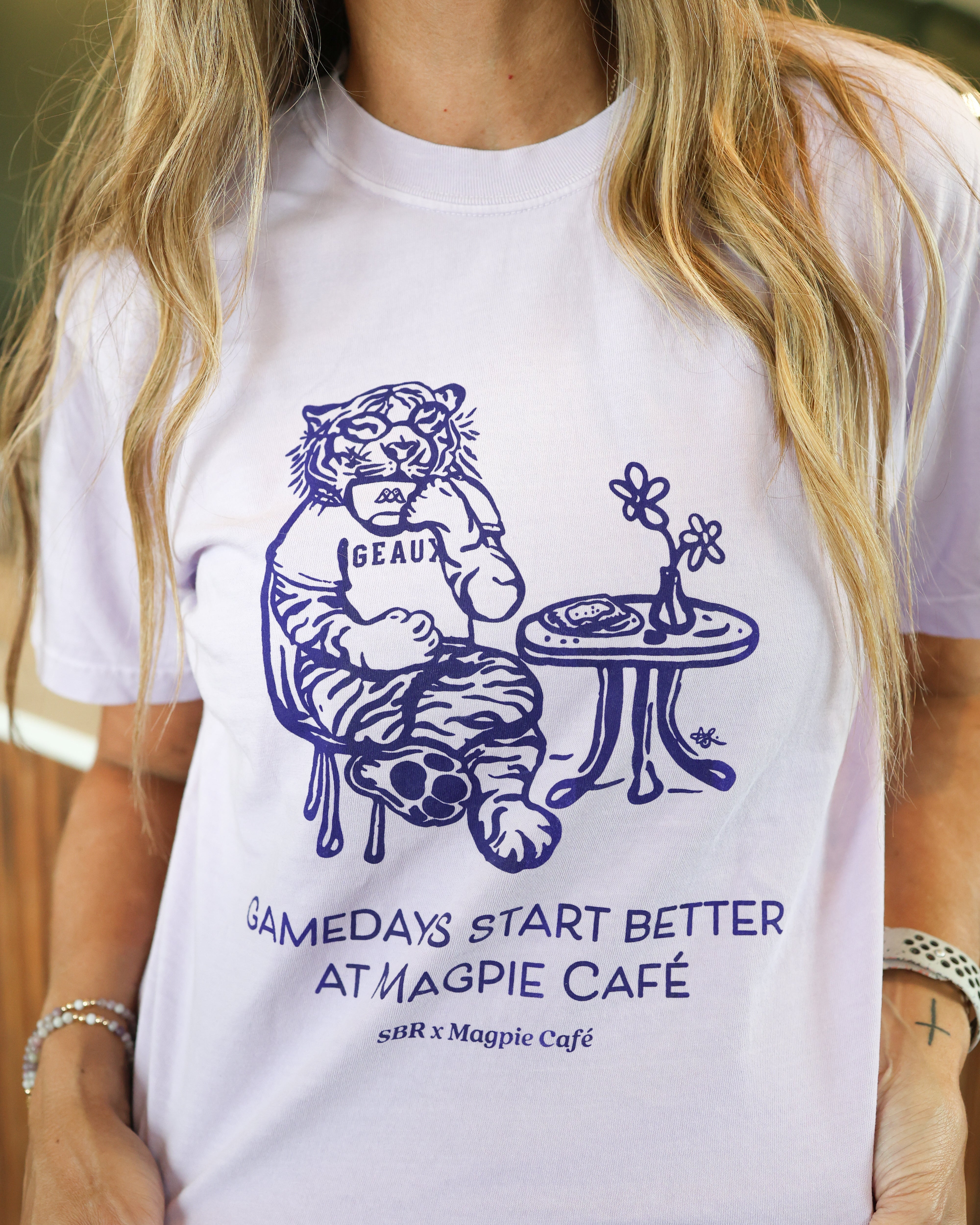 A woman orchid graphic t-shirt with a purple illustration of a tiger sitting at a table and the text 'GAMEDAYS START BETTER AT MAGPIE CAFÉ' and 'sR x Magpie Café'.