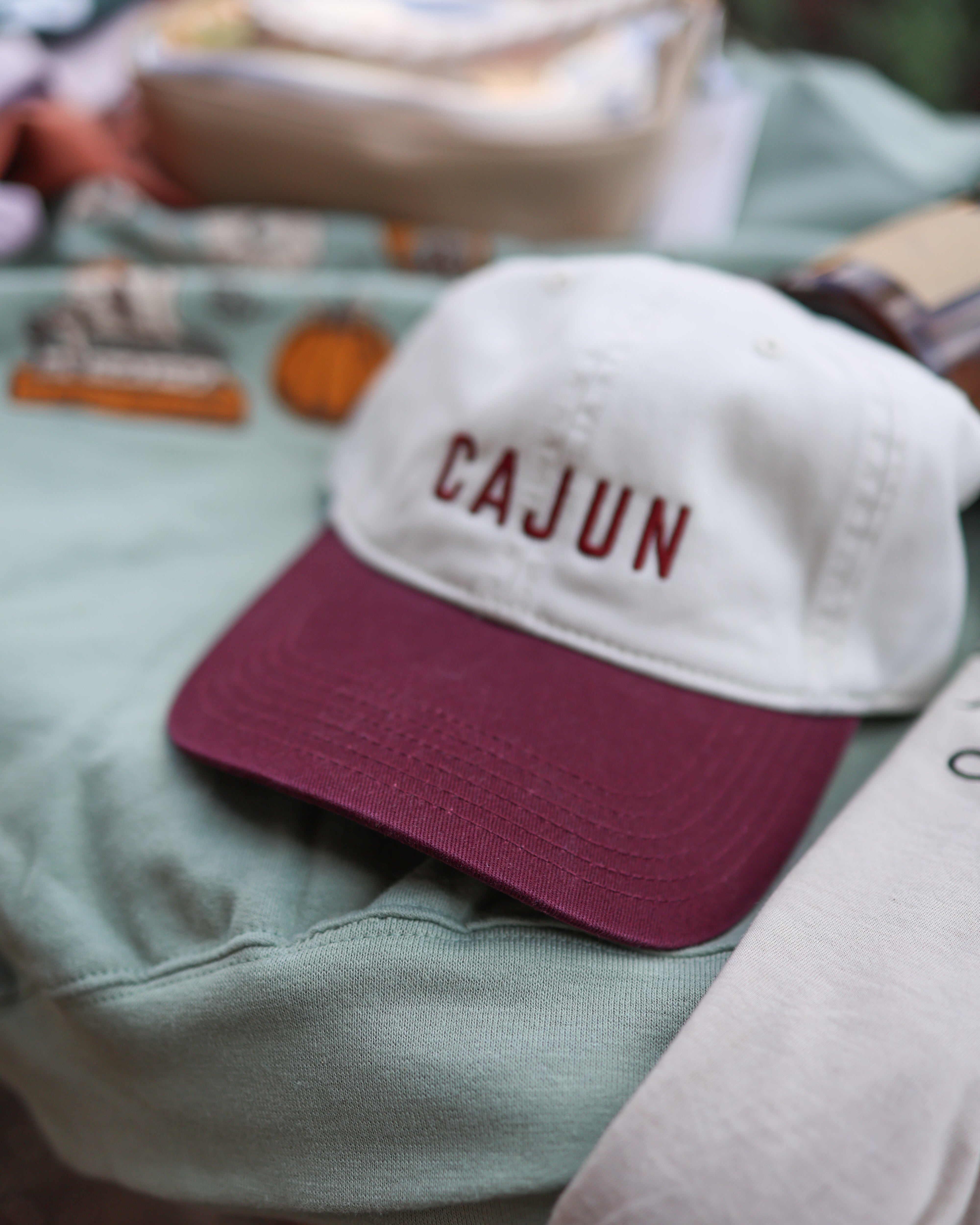 A white baseball cap with a burgundy brim and 'CAJUN' embroidered on the front, resting on a sage green sweatshirt.