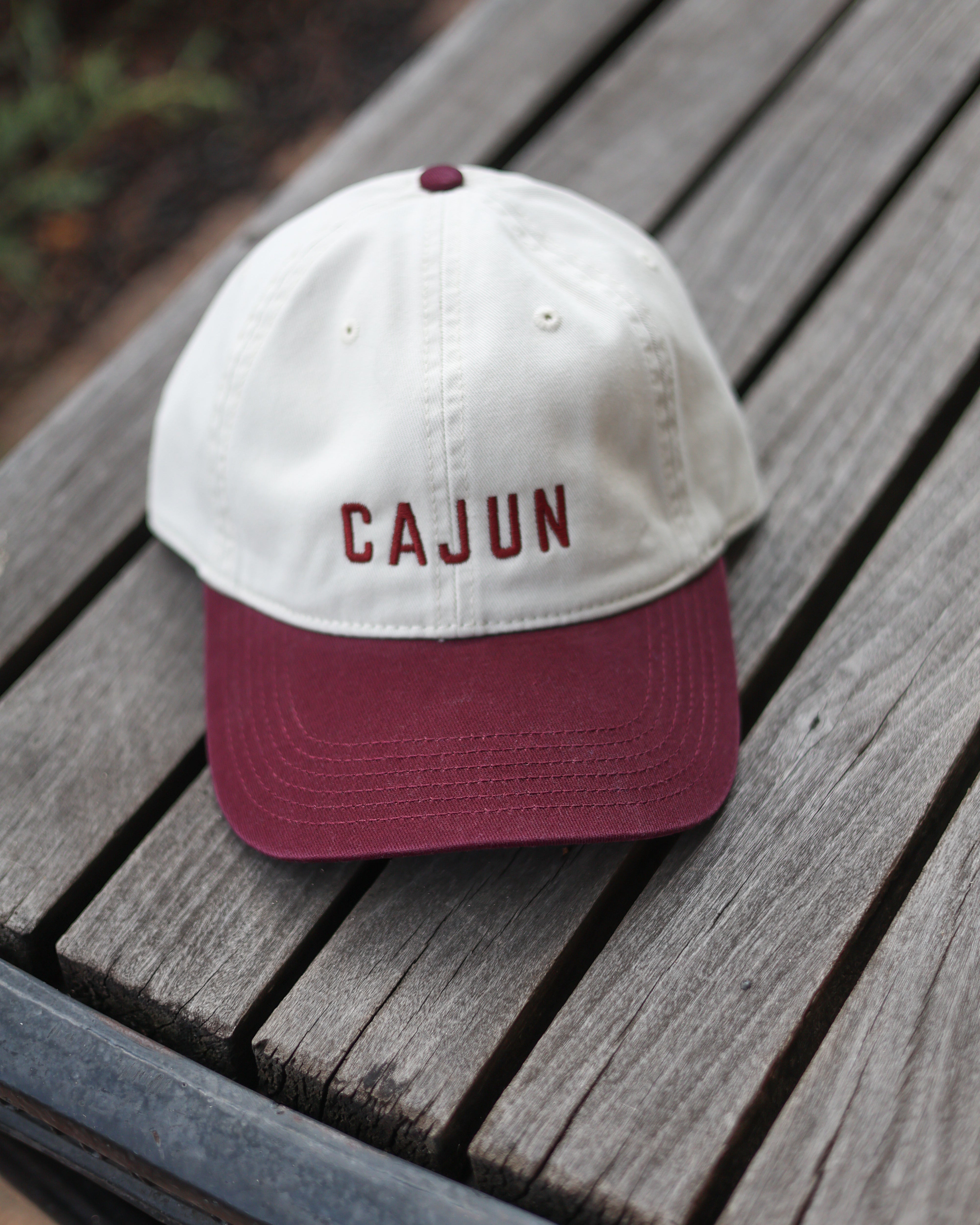 A two-tone baseball cap with an off-white crown and a maroon curved brim, featuring the word 'CAJUN' embroidered in maroon on the front. The cap is resting on weathered gray wooden slats.