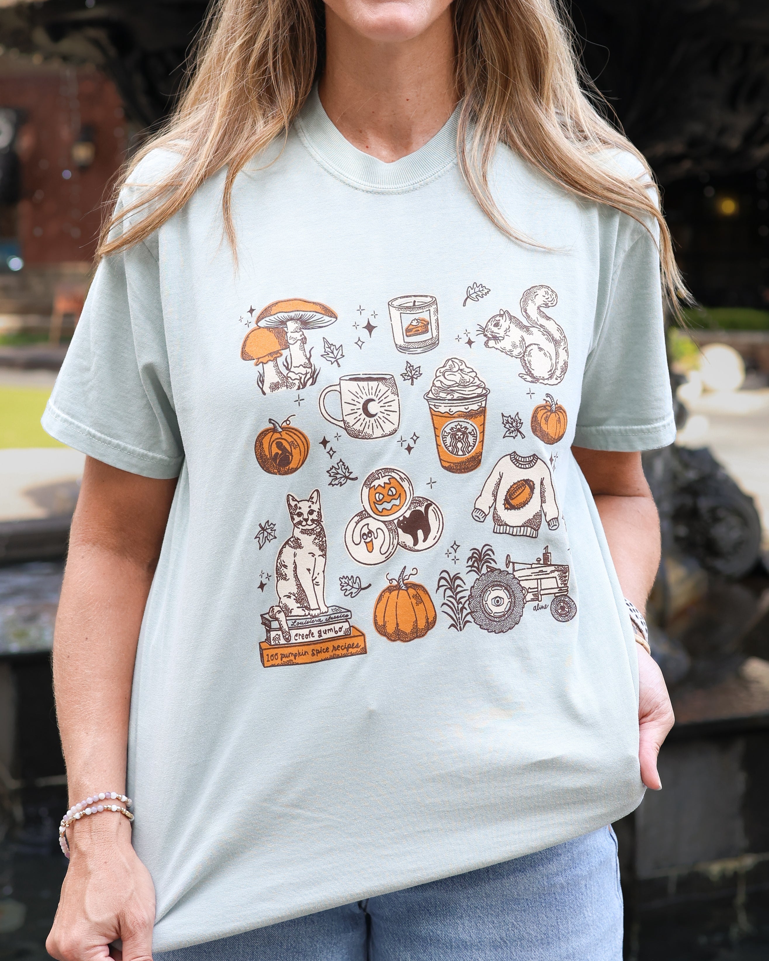 A woman wearing a light sage green graphic t-shirt with an autumn-themed print featuring pumpkins, a cat, a coffee cup, a squirrel, and a tractor.