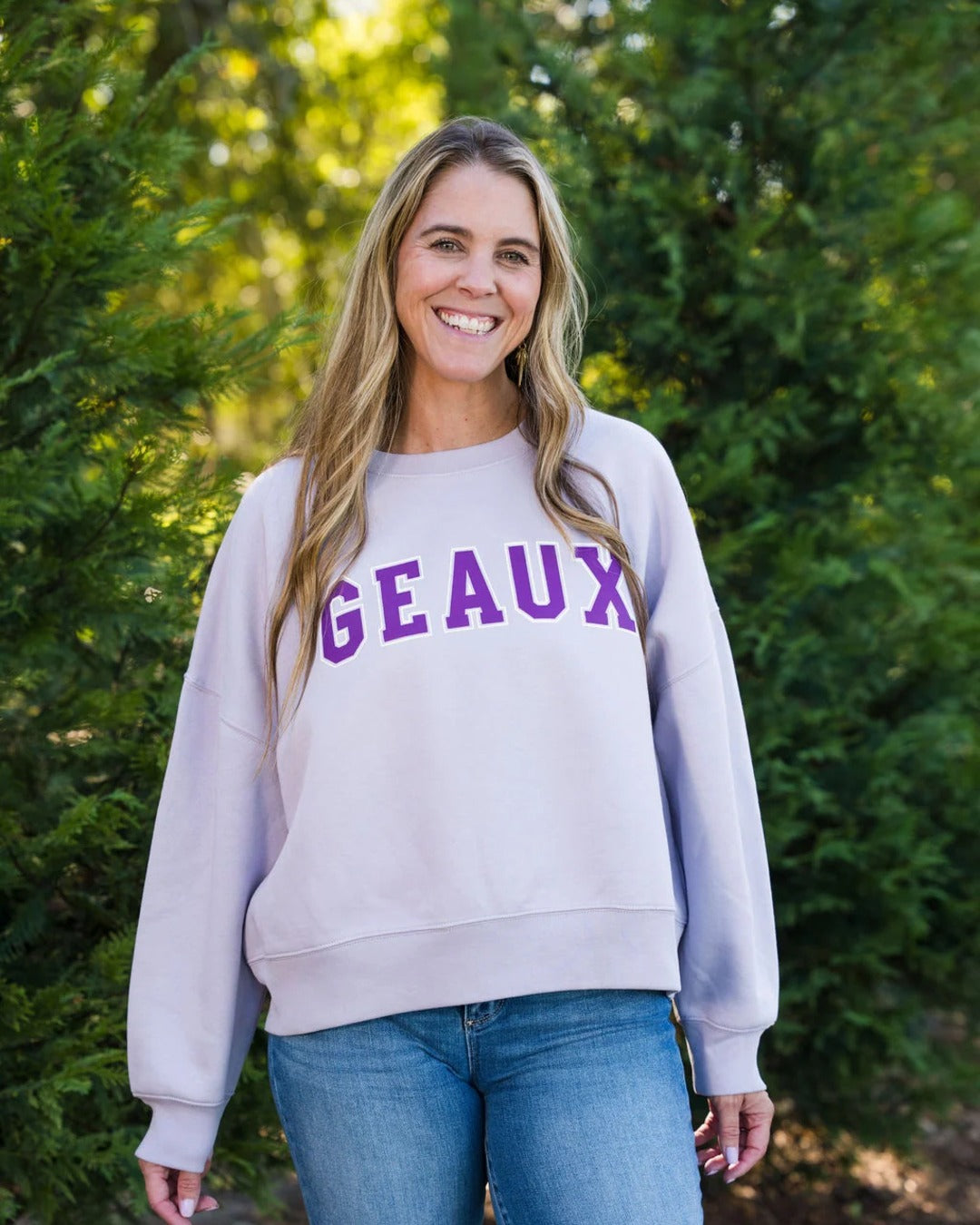 Smiling woman wearing the Geaux Block Cropped Sweatshirt in lavender with purple varsity GEAUX lettering and jeans