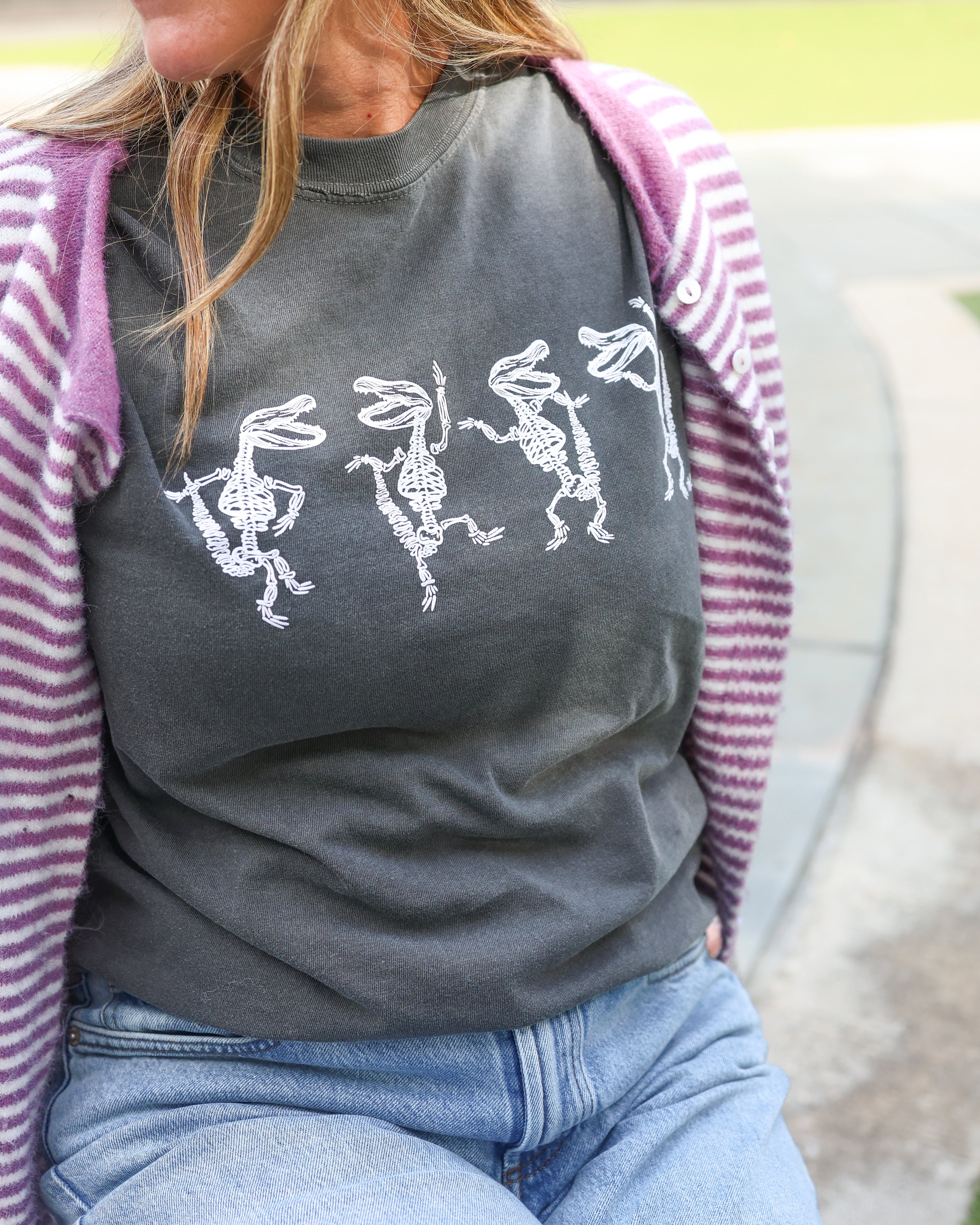 A woman wearing a dark gray t-shirt with a white graphic of four dancing figures, styled with a pink and white striped cardigan and light blue jeans.