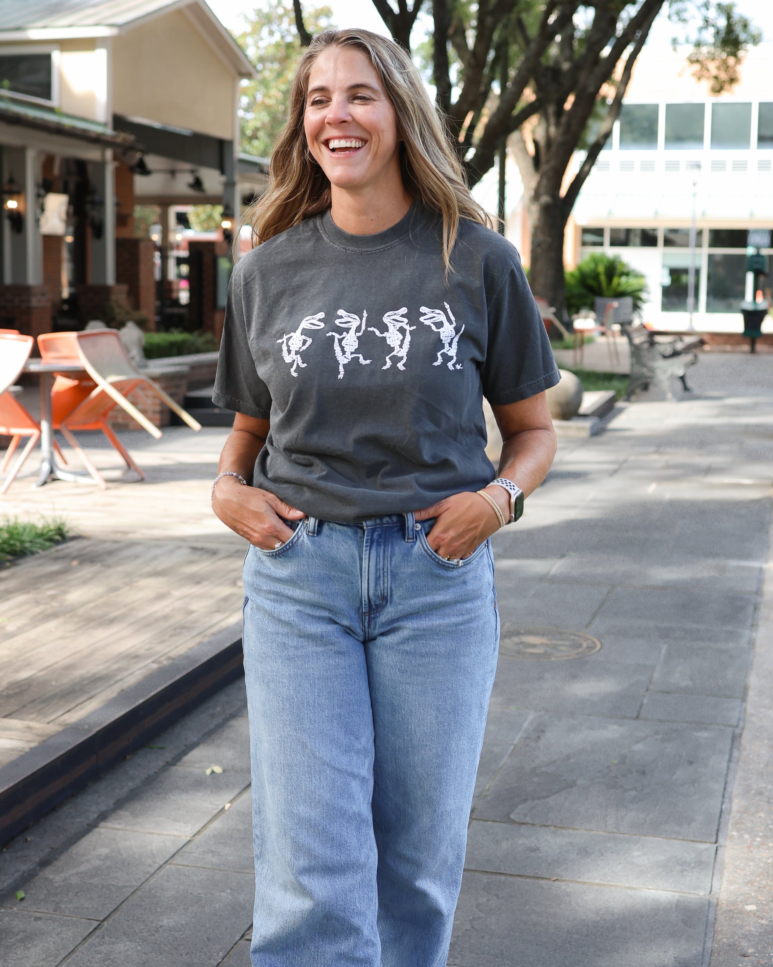 A woman walking outdoors on a paved path wearing a dark gray graphic t-shirt and light blue jeans.