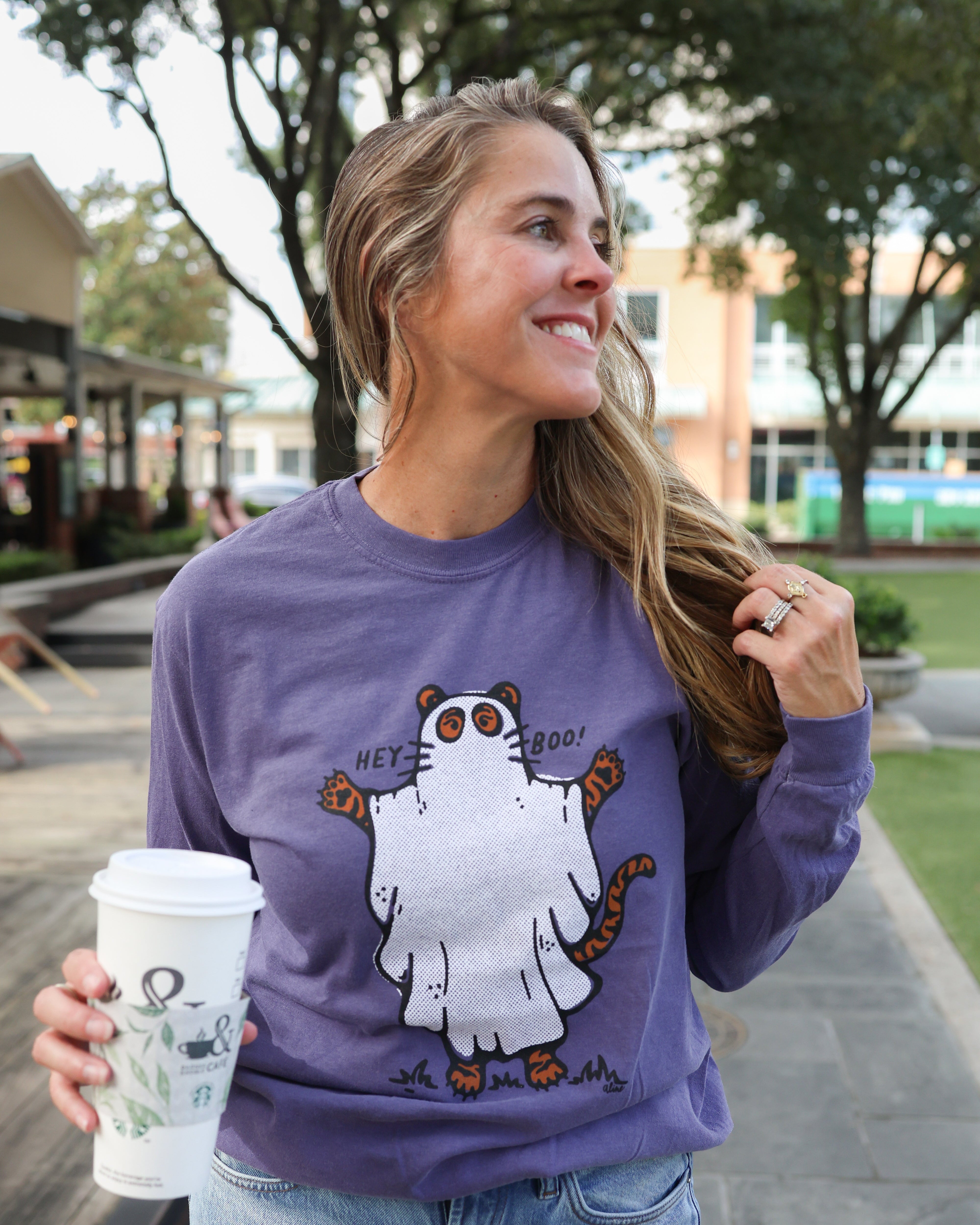 A woman wearing a purple crewneck sweatshirt with a cartoon ghost graphic that says 'Hey Boo!' stands outdoors on a paved walkway, holding a white coffee cup.