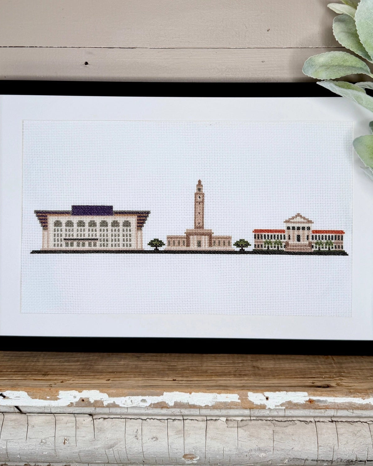 A framed art print featuring a colorful architectural LSU illustration, styled on a rustic wooden shelf next to a white ceramic vase with green leaves against a beige wood-paneled wall.