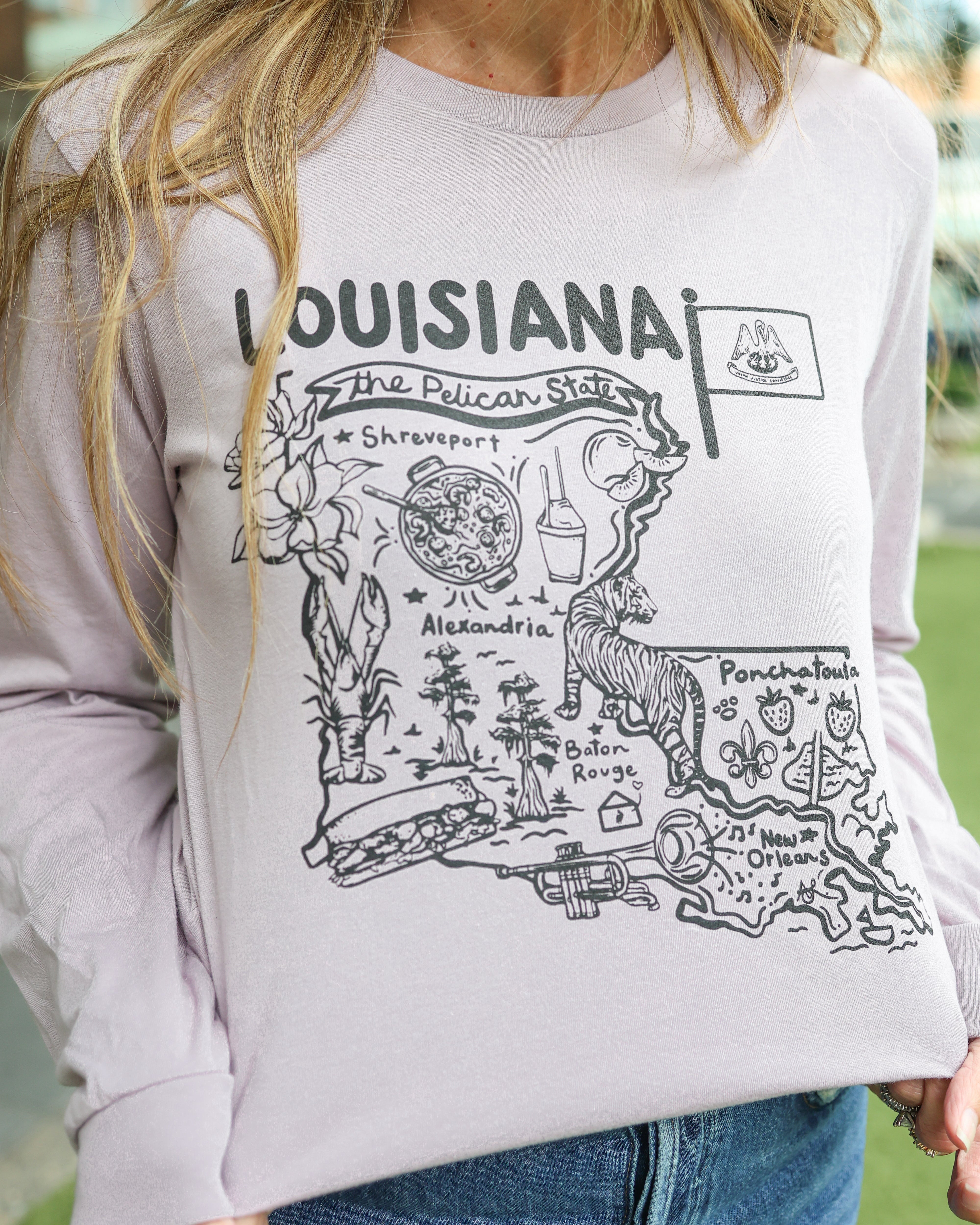 A woman wearing a light lavender long-sleeve crewneck sweatshirt with a black graphic print of the Louisiana state outline and 'Louisiana The Pelican State' text, paired with blue jeans.