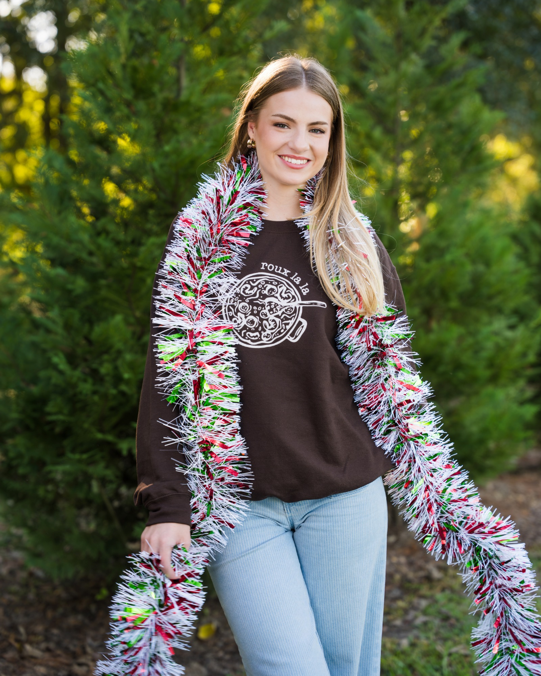 Smiling woman wearing the dark chocolate Roux La La Sweatshirt from the Gumbo Roux La La Gift Set, styled with a festive tinsel boa