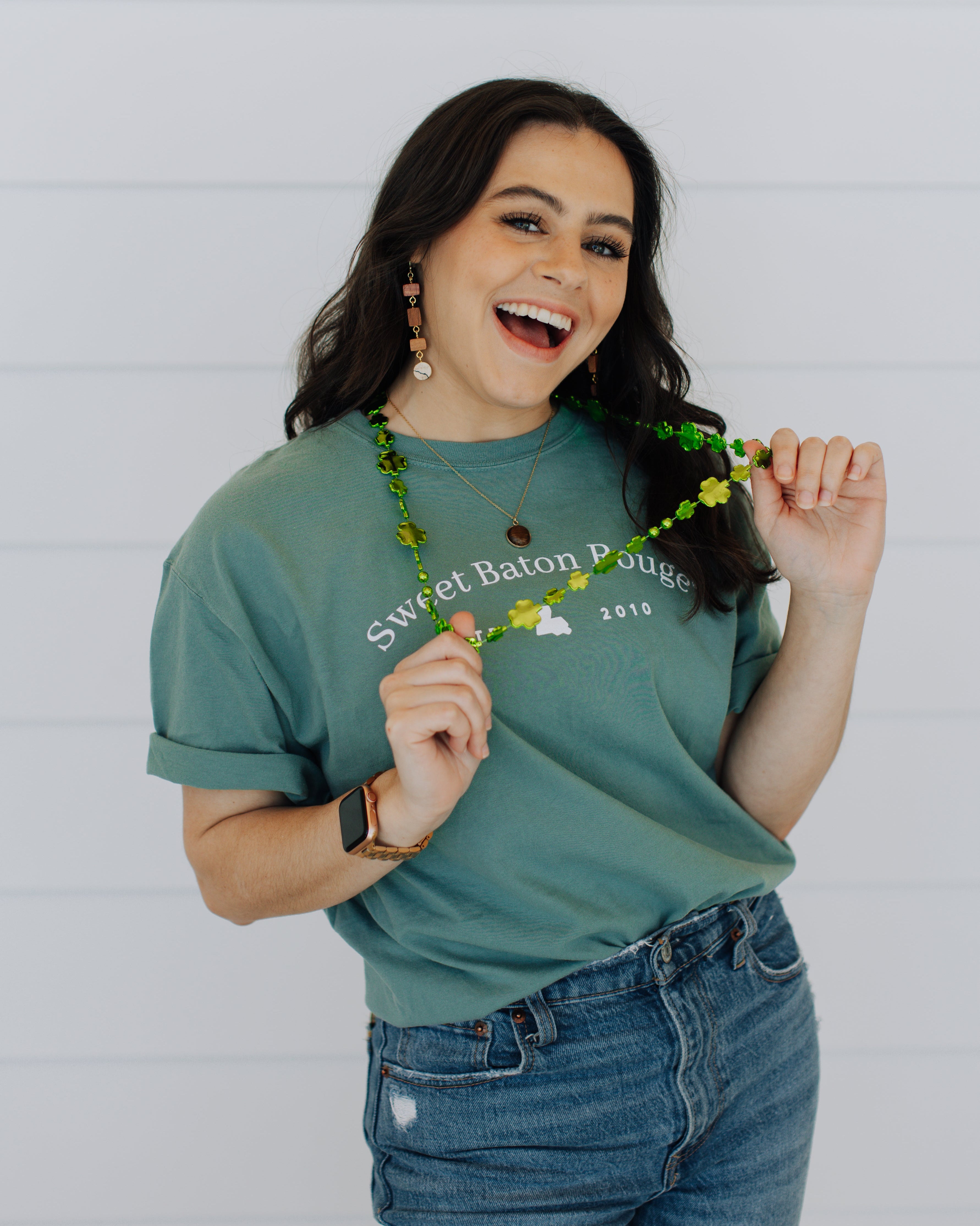 Woman wearing a green Sweet Baton Rouge® t-shirt and blue jeans, holding a decorative item against a white background
