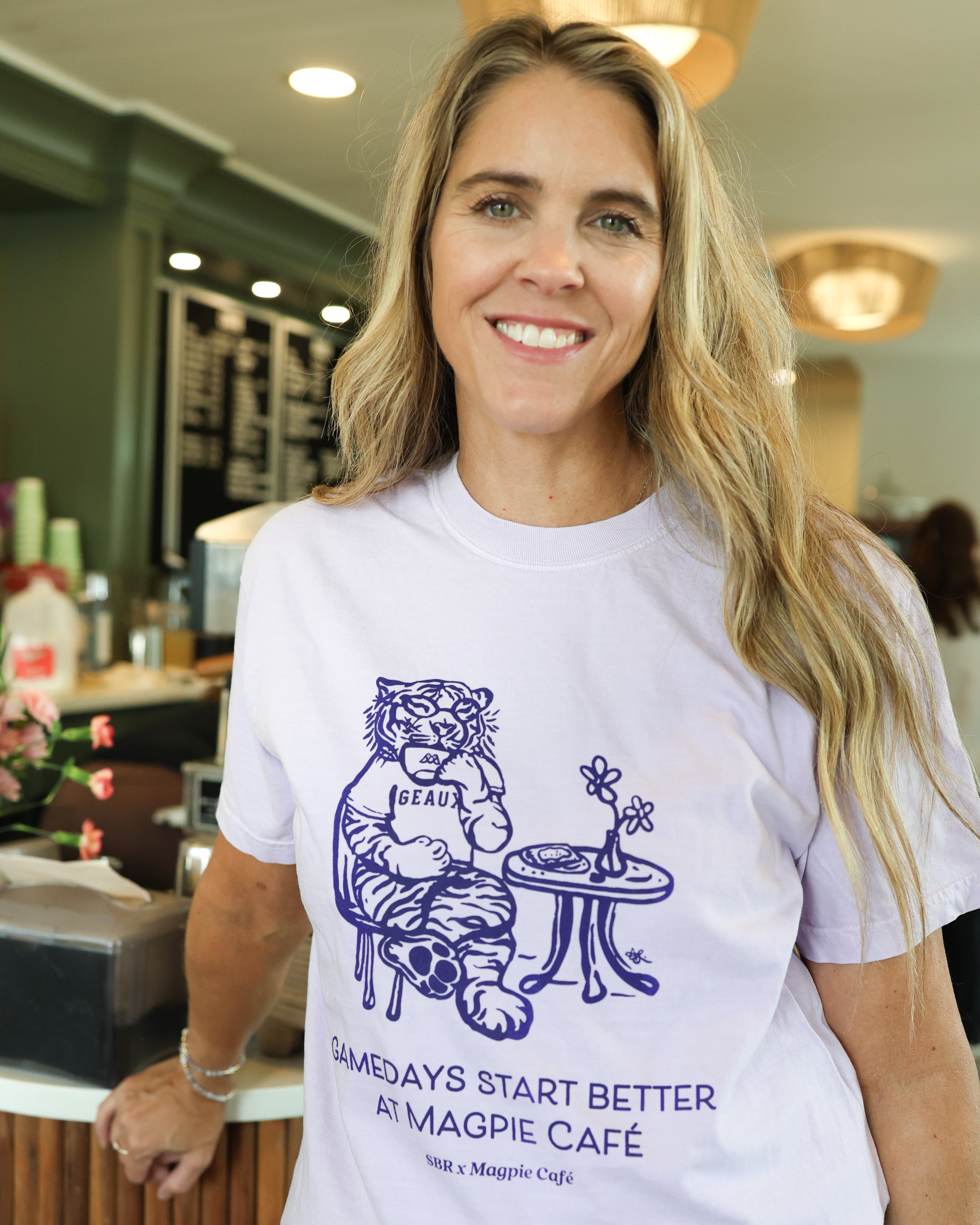 A smiling woman with blonde hair stands in a cafe wearing a white crewneck t-shirt with a dark purple graphic of a tiger sitting at a table and the text 'GAMEDAYS START BETTER AT MAGPIE CAFE'.