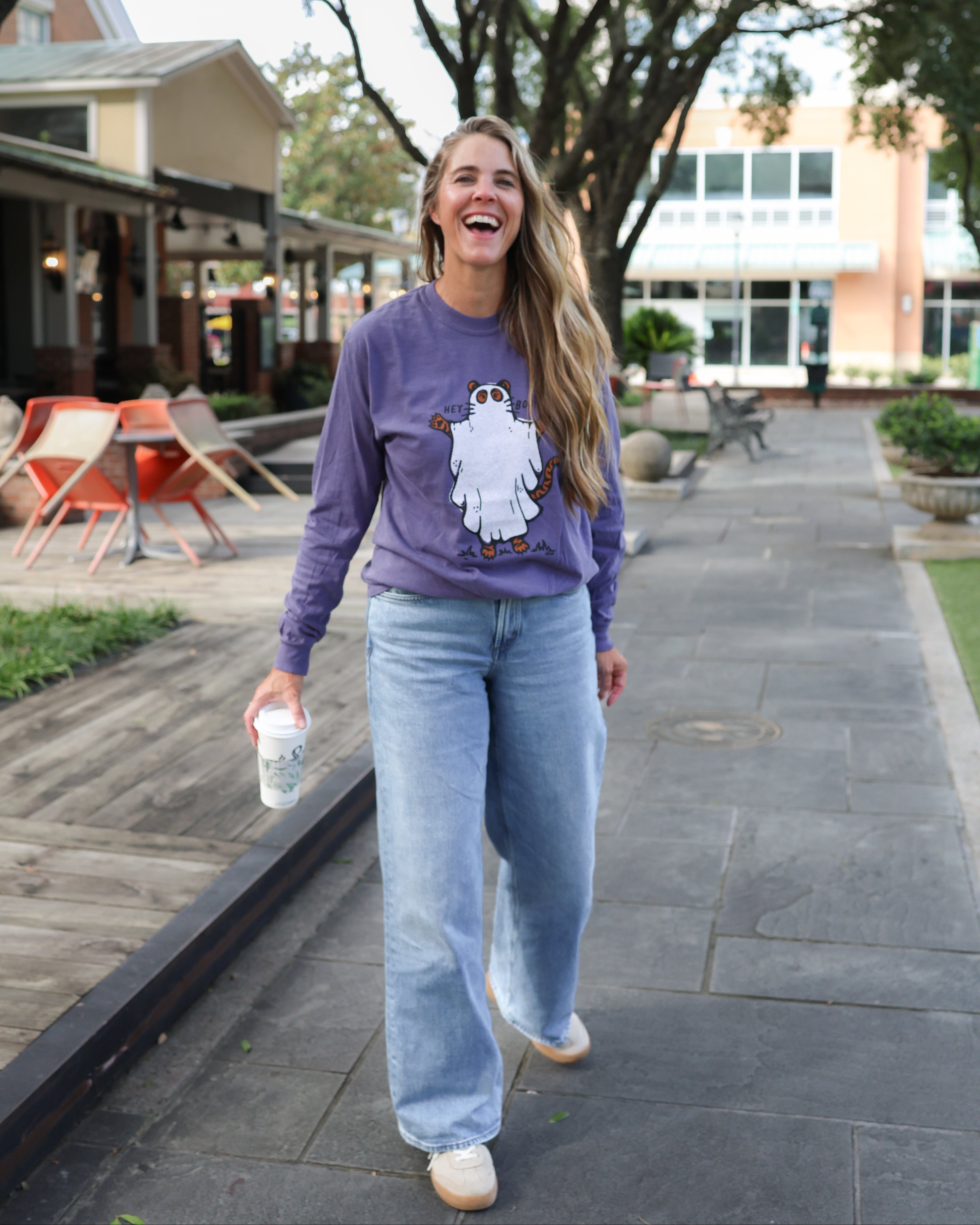A woman laughing while walking outdoors on a paved path, wearing a purple long-sleeve sweatshirt with a white cartoon tiger ghost graphic on the front, light blue jeans, and white sneakers.