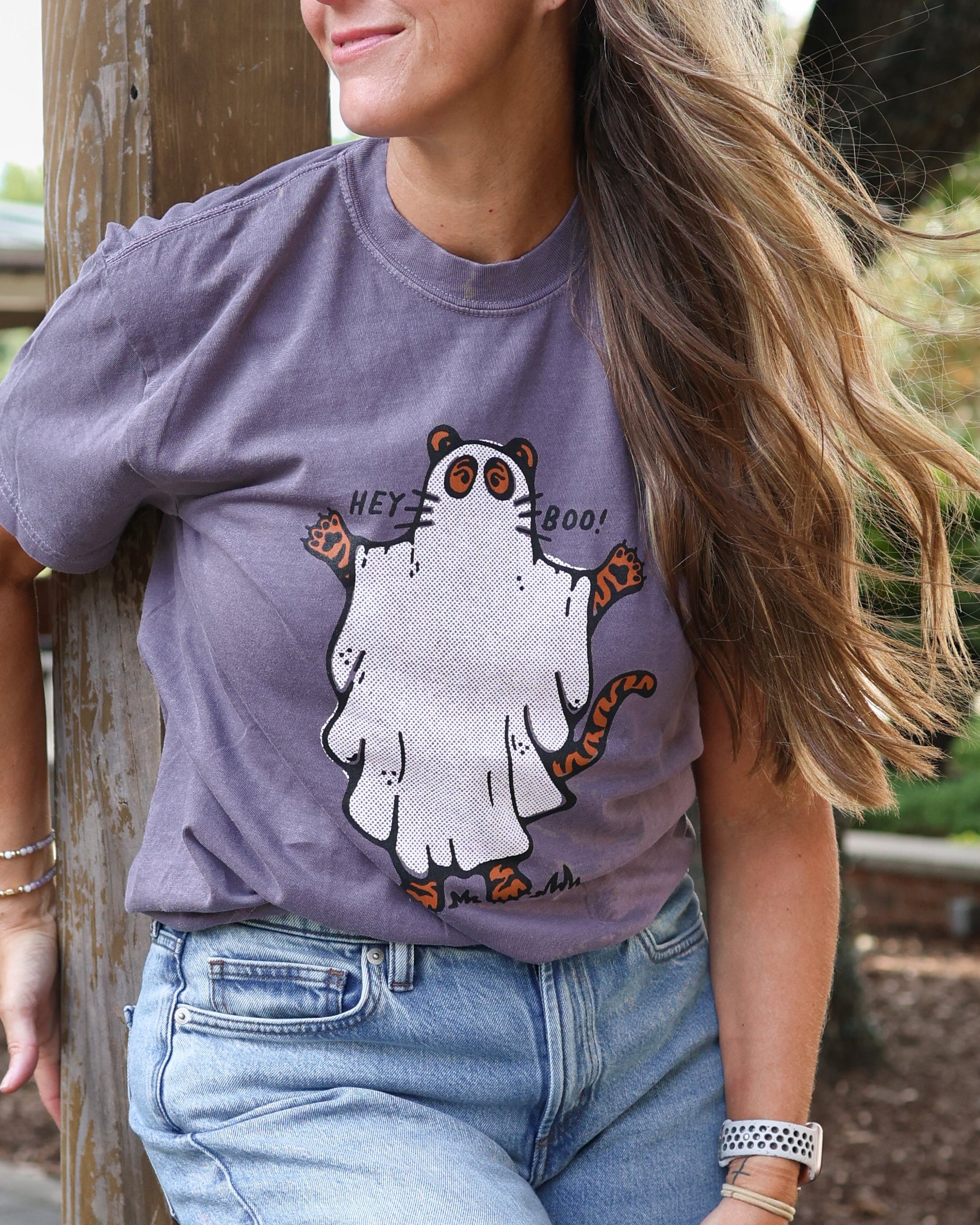 A woman with long light brown hair leans against a wooden post on an outdoor deck, wearing a purple graphic t-shirt with a cartoon ghost and the text 'Hey Boo' and light wash blue jeans.