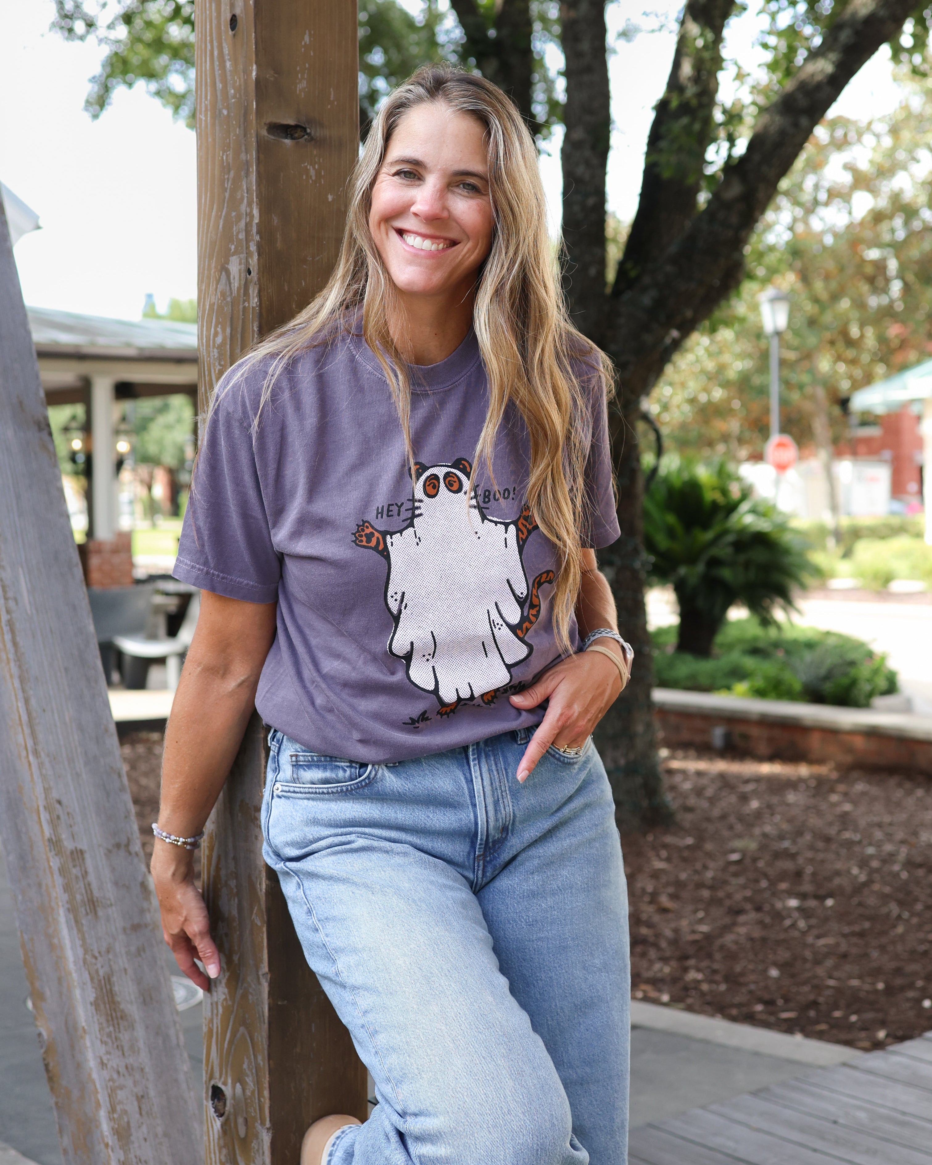 A woman smiling and leaning against a wooden post on an outdoor deck, wearing a purple t-shirt with a white ghost graphic and light-wash blue jeans.