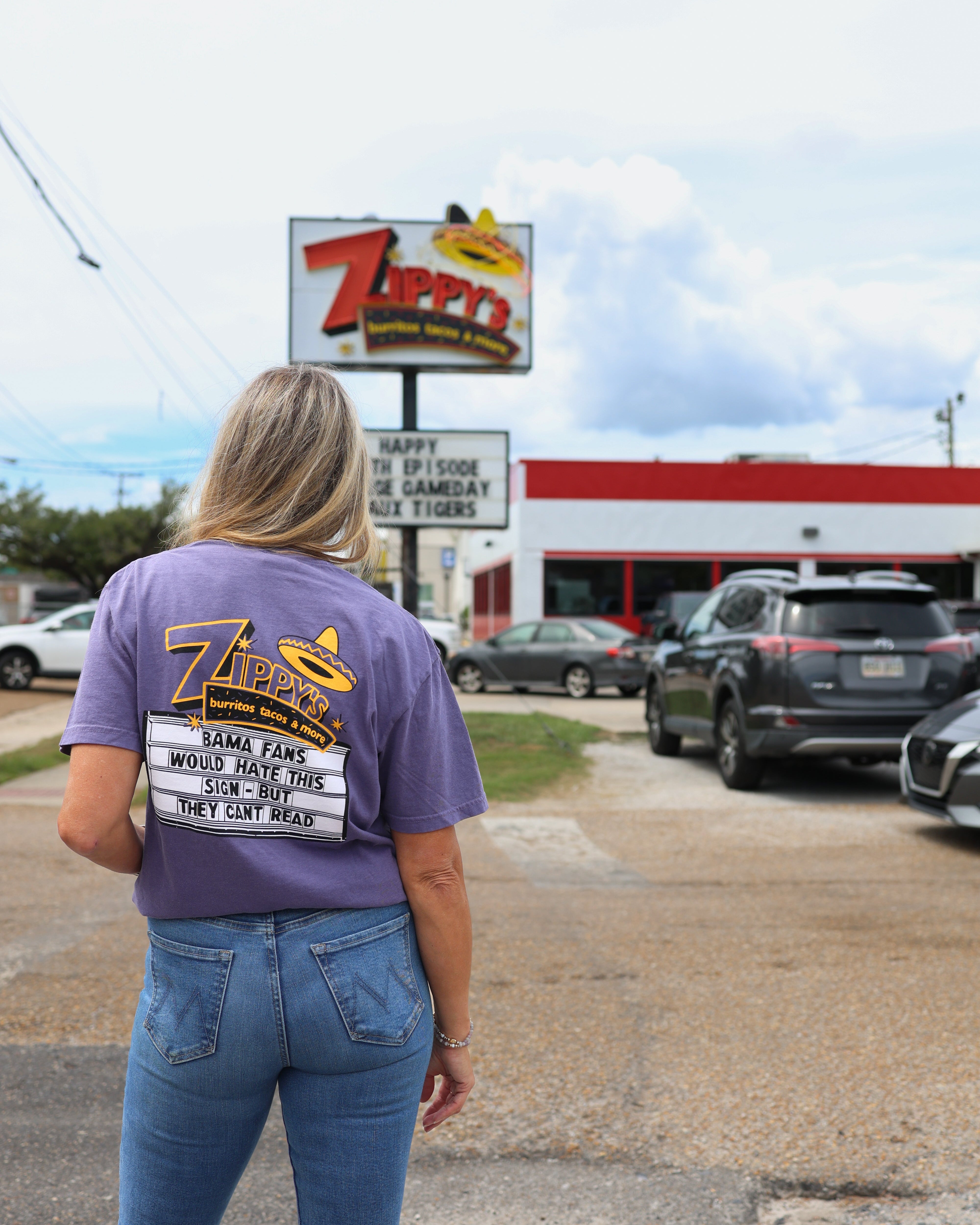 A woman viewed from behind wearing a purple graphic t-shirt with a 'Zippy's' restaurant logo, standing in a gravel lot in front of the restaurant building and sign.
