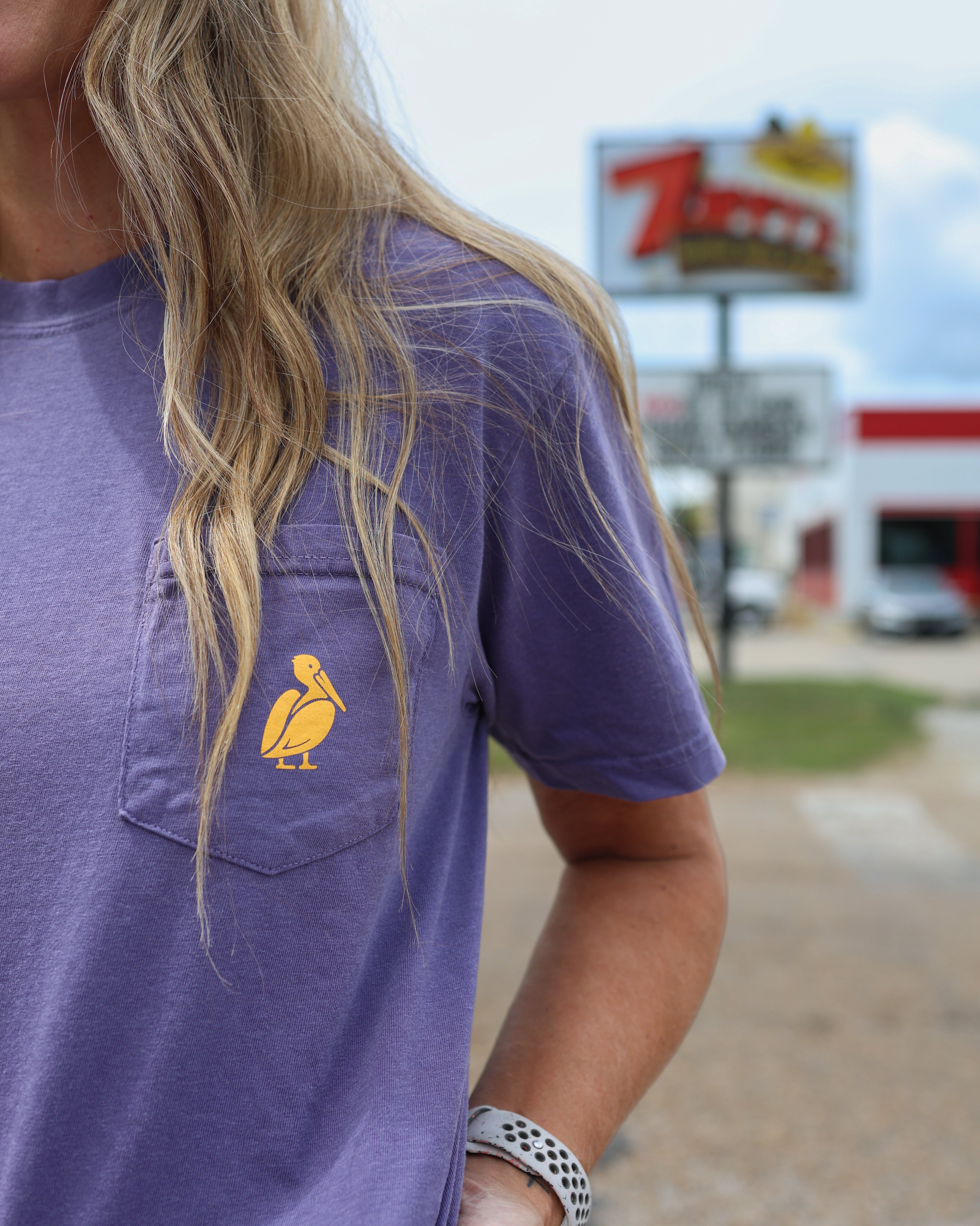 A woman wearing a heathered purple short-sleeve pocket t-shirt with a yellow pelican logo on the chest, photographed outdoors with a blurred background.
